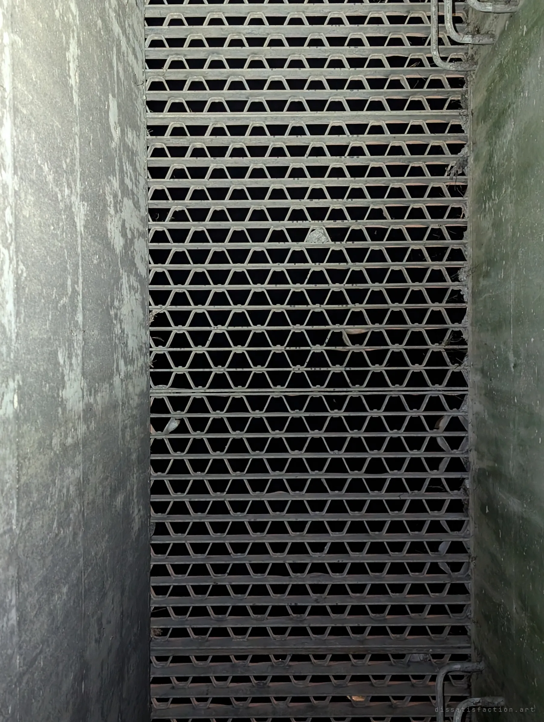 Looking up a narrow concrete shaft at a steel grating with a repeating hexagon pattern, ladder rungs at top and bottom. The grate lets the drain breathe to the surface.