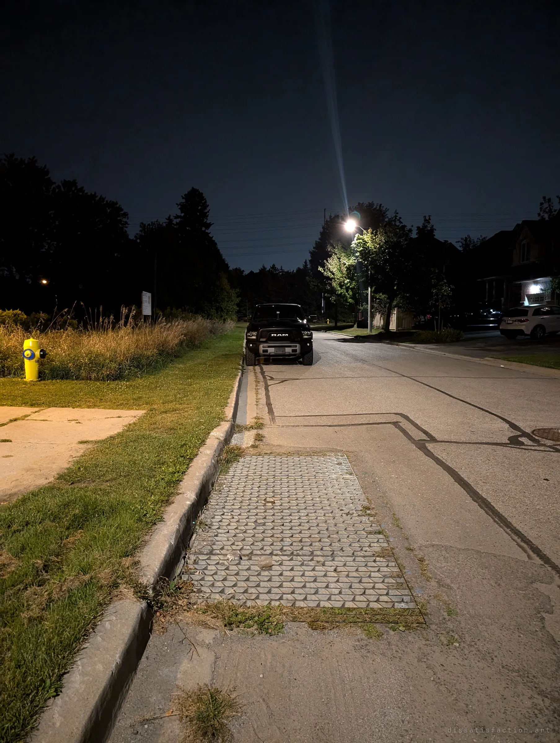 The drain's curbside grate seen from the street at night, set into the gutter of a quiet residential road under a single streetlight. It shows how ordinary the way in looks from above.