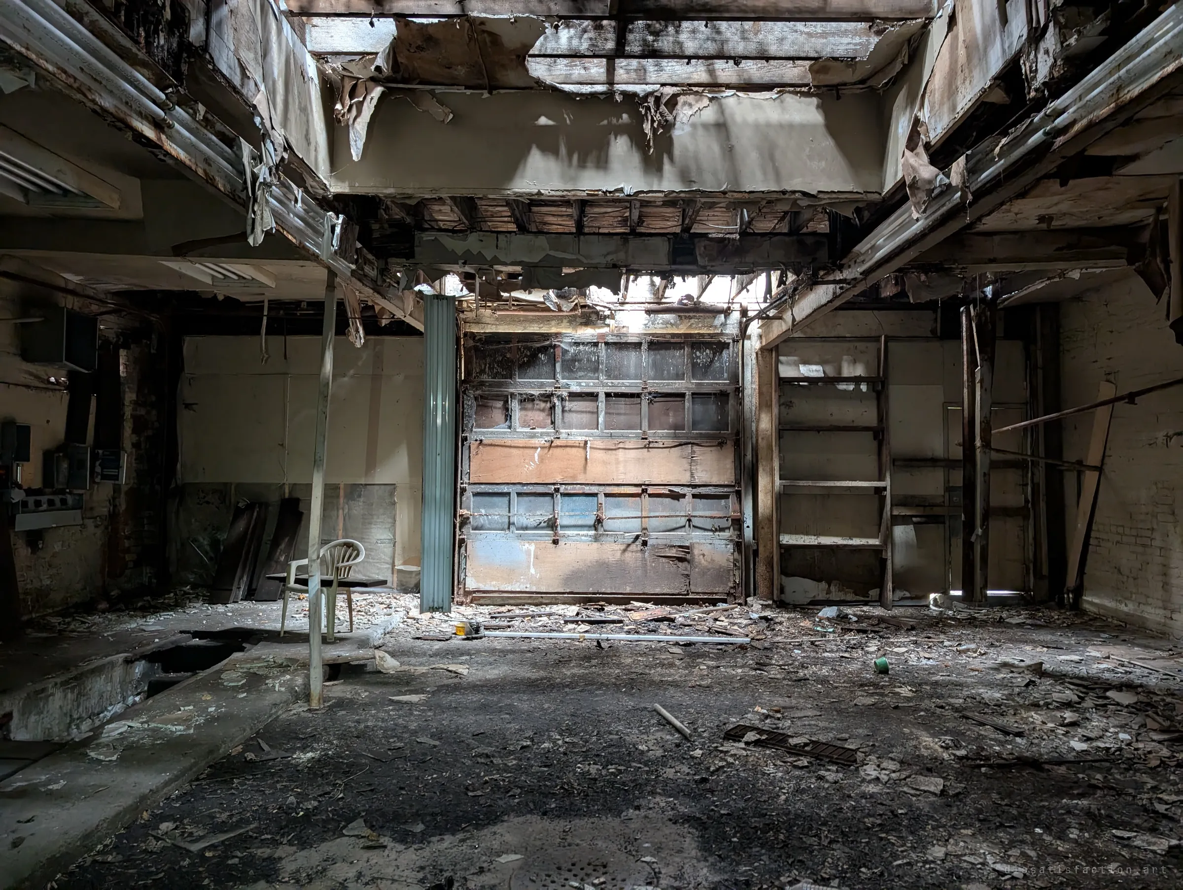 Daylight pours through a collapsed ceiling onto a large overhead door, a lone plastic chair and a floor thick with debris. The open roof shows how far the decay has gone.