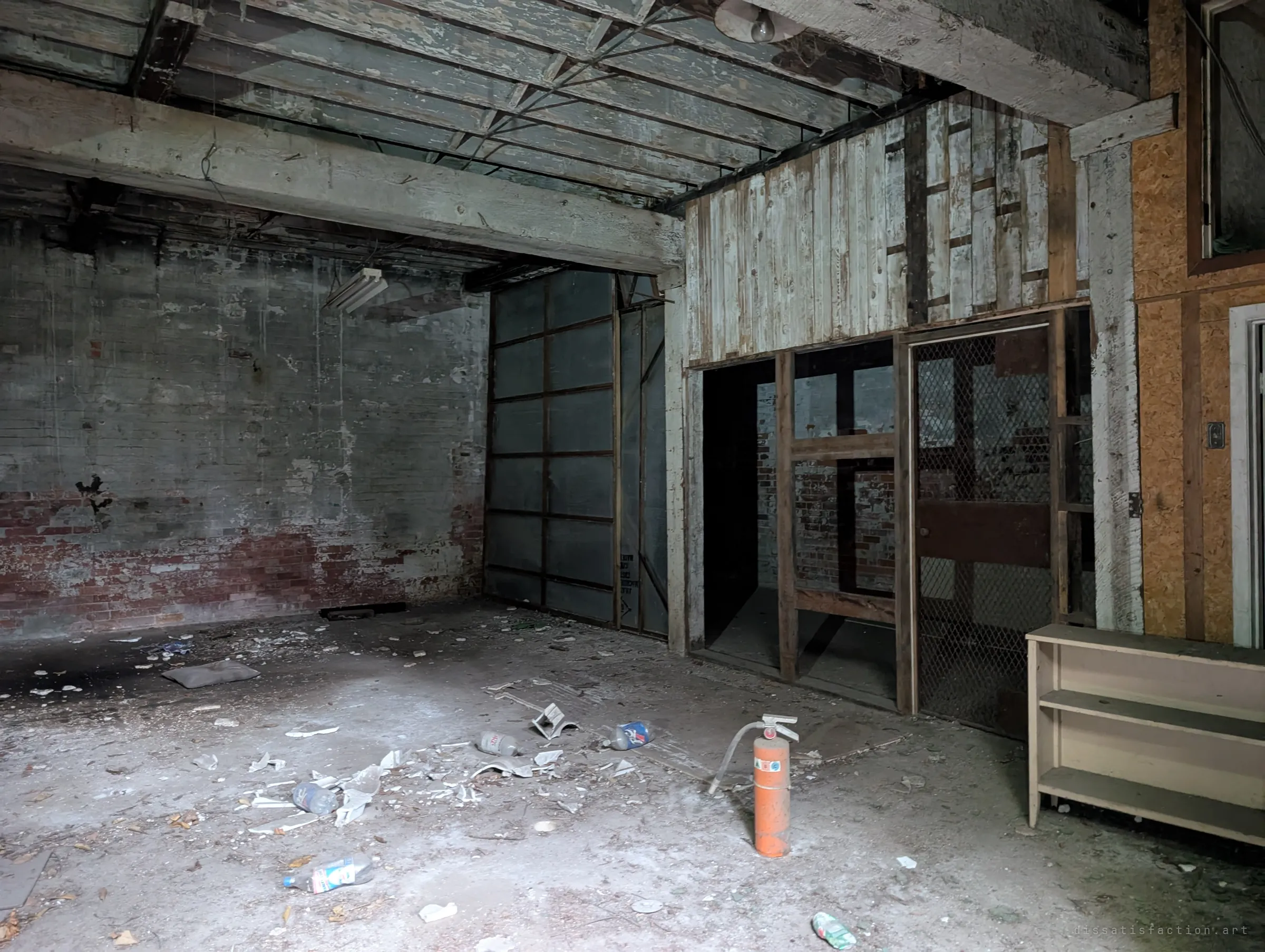 A dim storage room with whitewashed brick, a timber beam ceiling and a wire-mesh cage, with an old fire extinguisher standing among bottles on the floor.