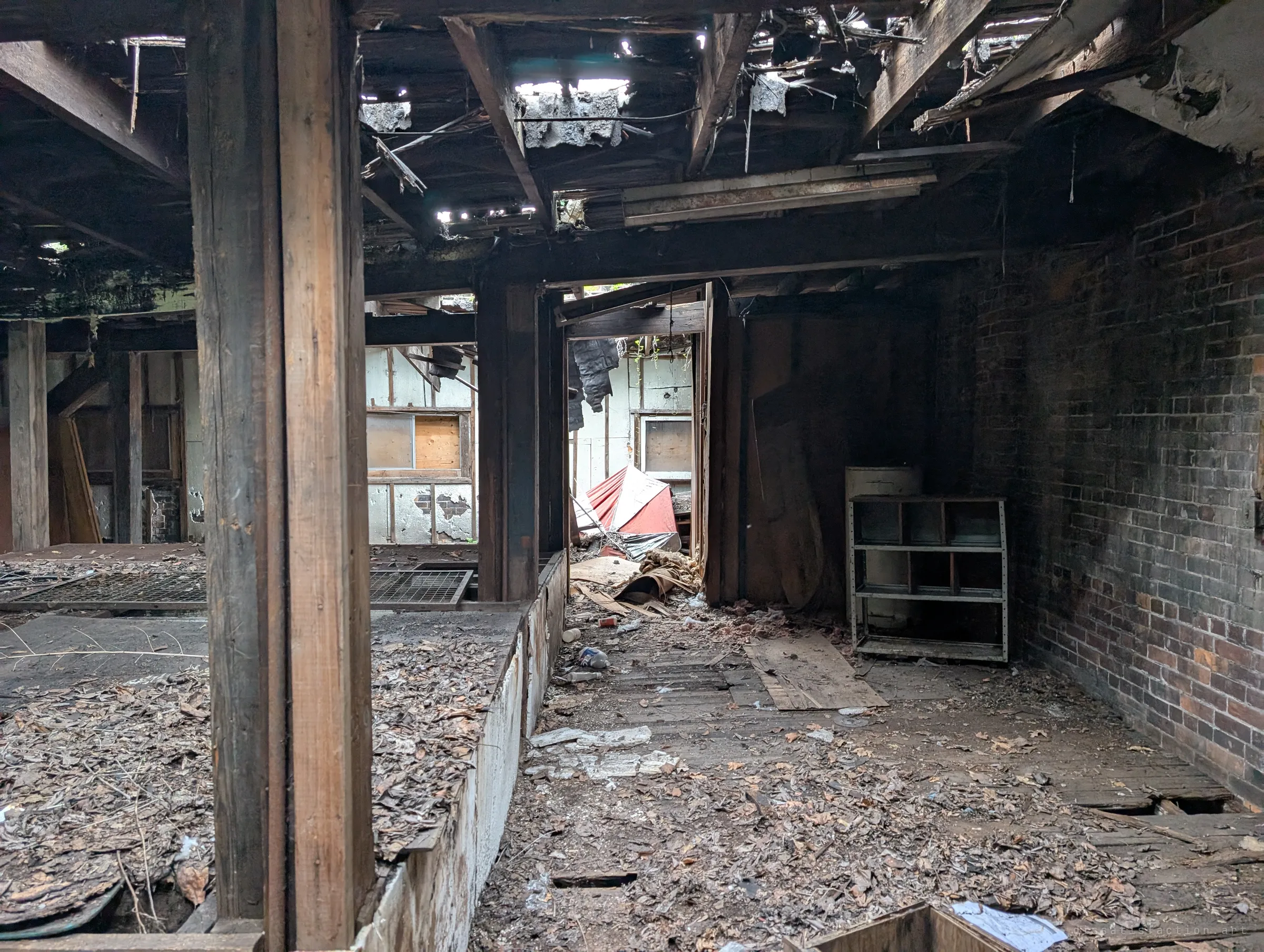 A leaf-strewn corridor runs between the raised beds and a soot-darkened brick wall toward a collapsed red and white umbrella, with light leaking through holes in the ceiling.