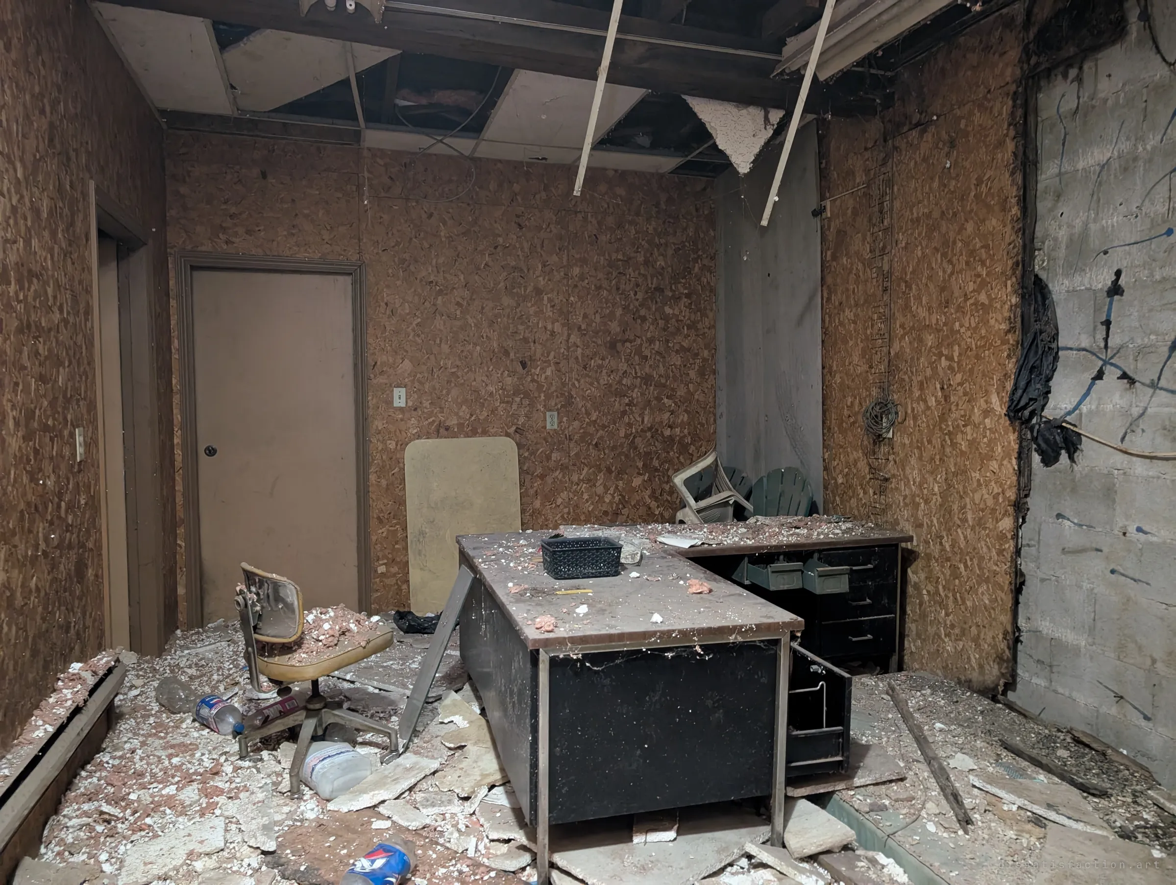 A small office with chipboard walls and a caved-in drop ceiling, its metal desk and chair covered in fallen insulation and plaster. The paperwork is long gone but the furniture stayed.