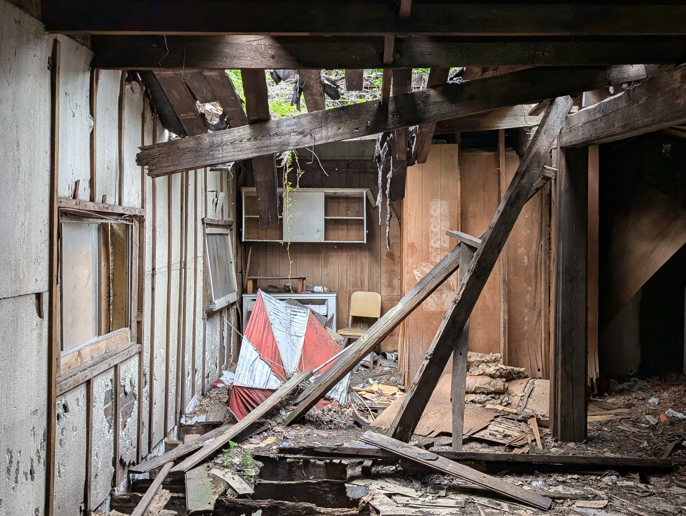 Broken roof beams lean across a back room where plants creep in through the open ceiling, over a crumpled red and white umbrella and an abandoned yellow chair.