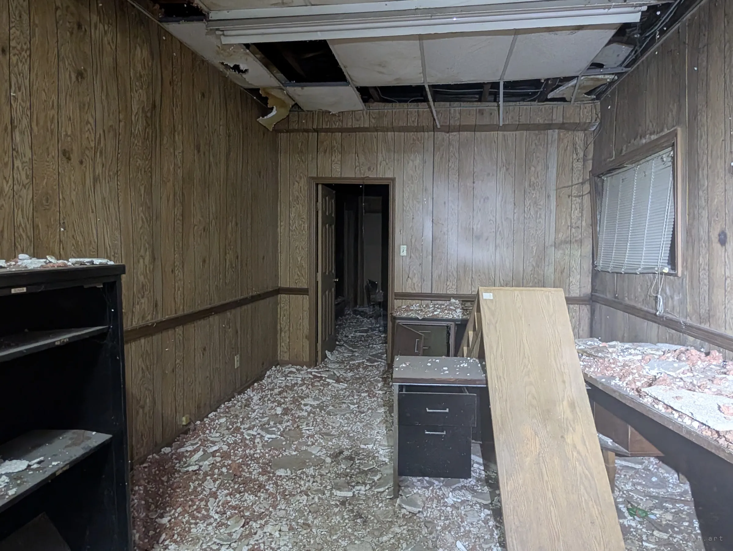 A wood-panelled back office buried in fallen ceiling tiles, with an open door leading into a dark hallway. The collapsed drop ceiling and abandoned desks show how long the building has sat empty.