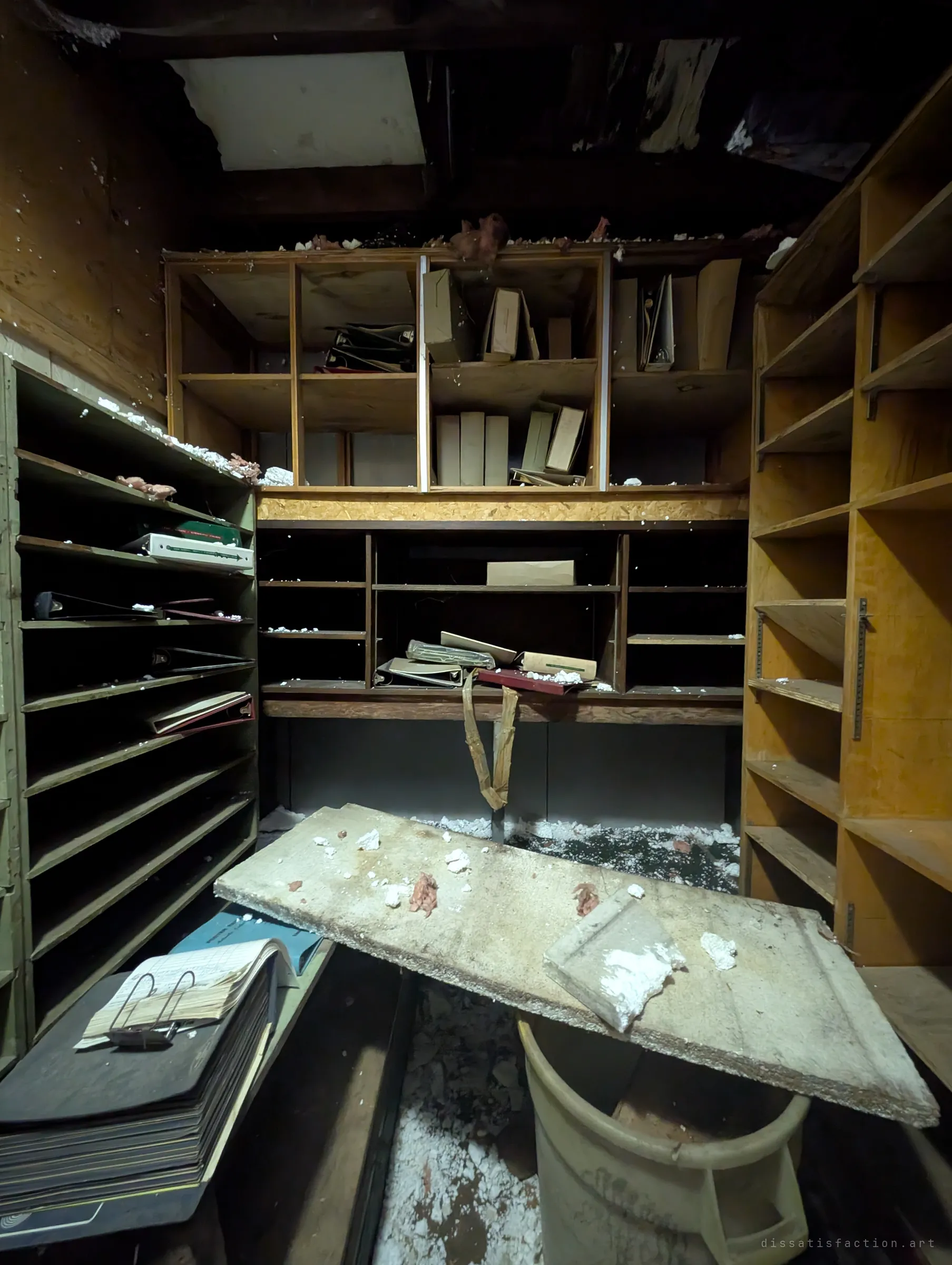 A cramped storeroom of pigeonhole shelving still holding old binders and a clipped ledger, with a broken panel resting across a bin. The paperwork left behind hints at a business that closed in a hurry.