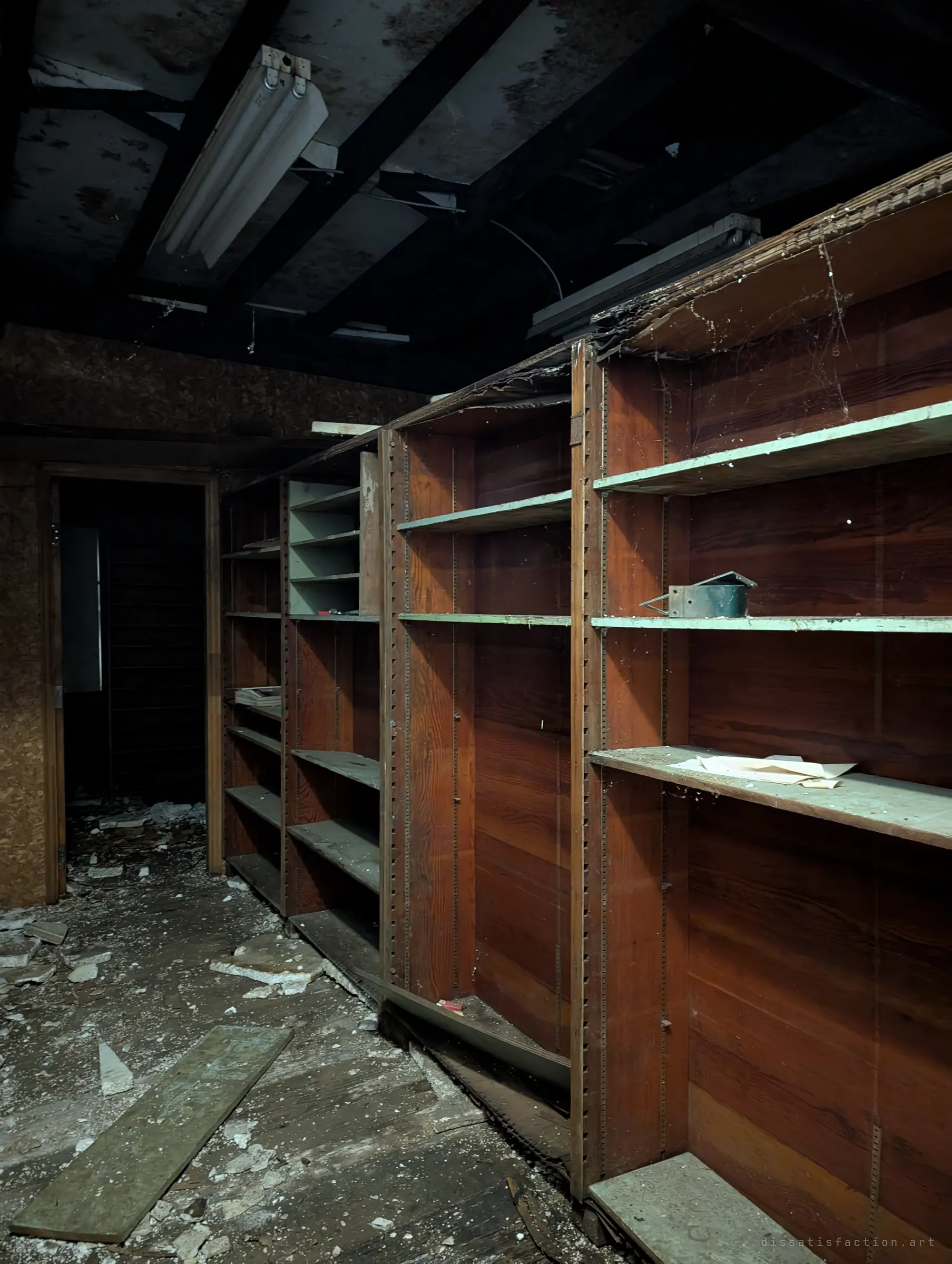 Rows of tall wooden shelving units stand empty along a dark stockroom under a blackened ceiling. A few loose papers and a small metal tray are all that remain of the stock.