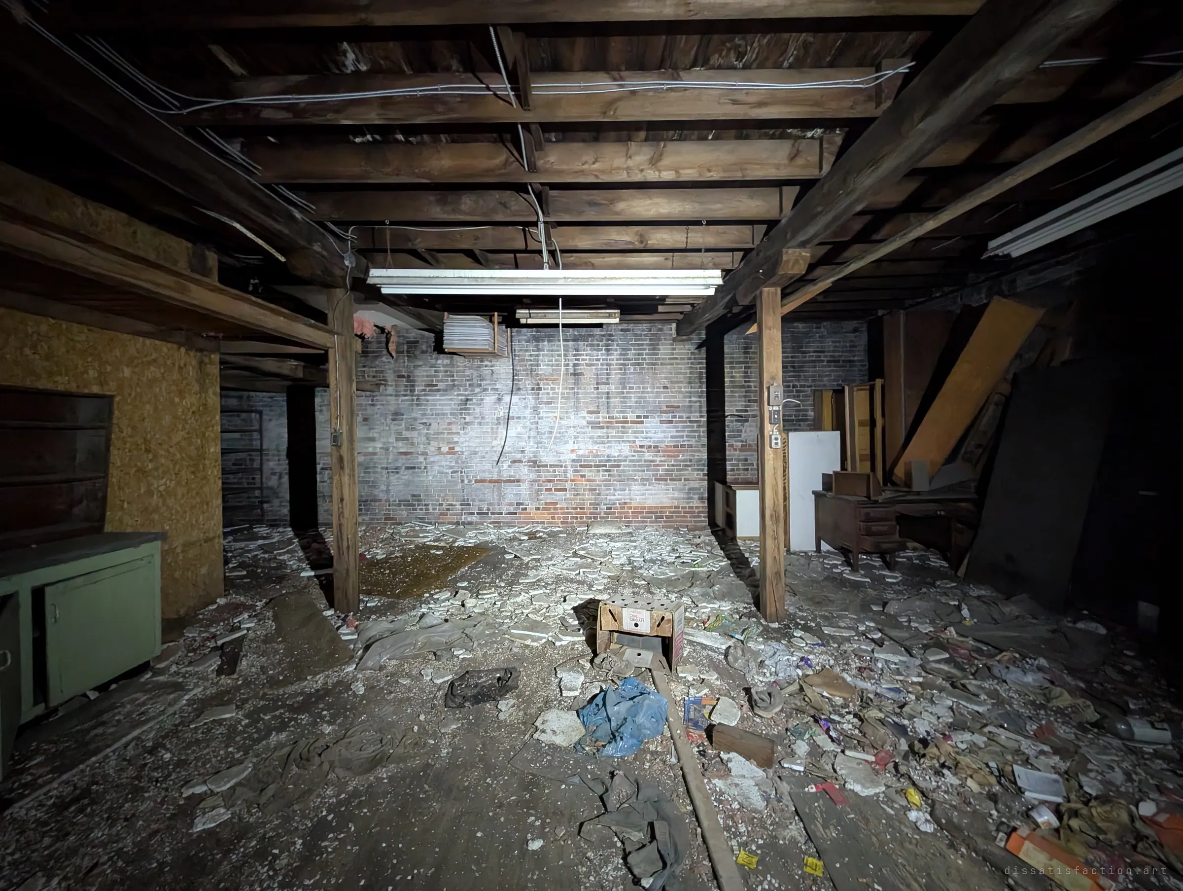 A low basement with heavy timber joists and posts, a whitewashed brick back wall and a floor strewn with debris and trash. A single work light picks out the old structure of the building.