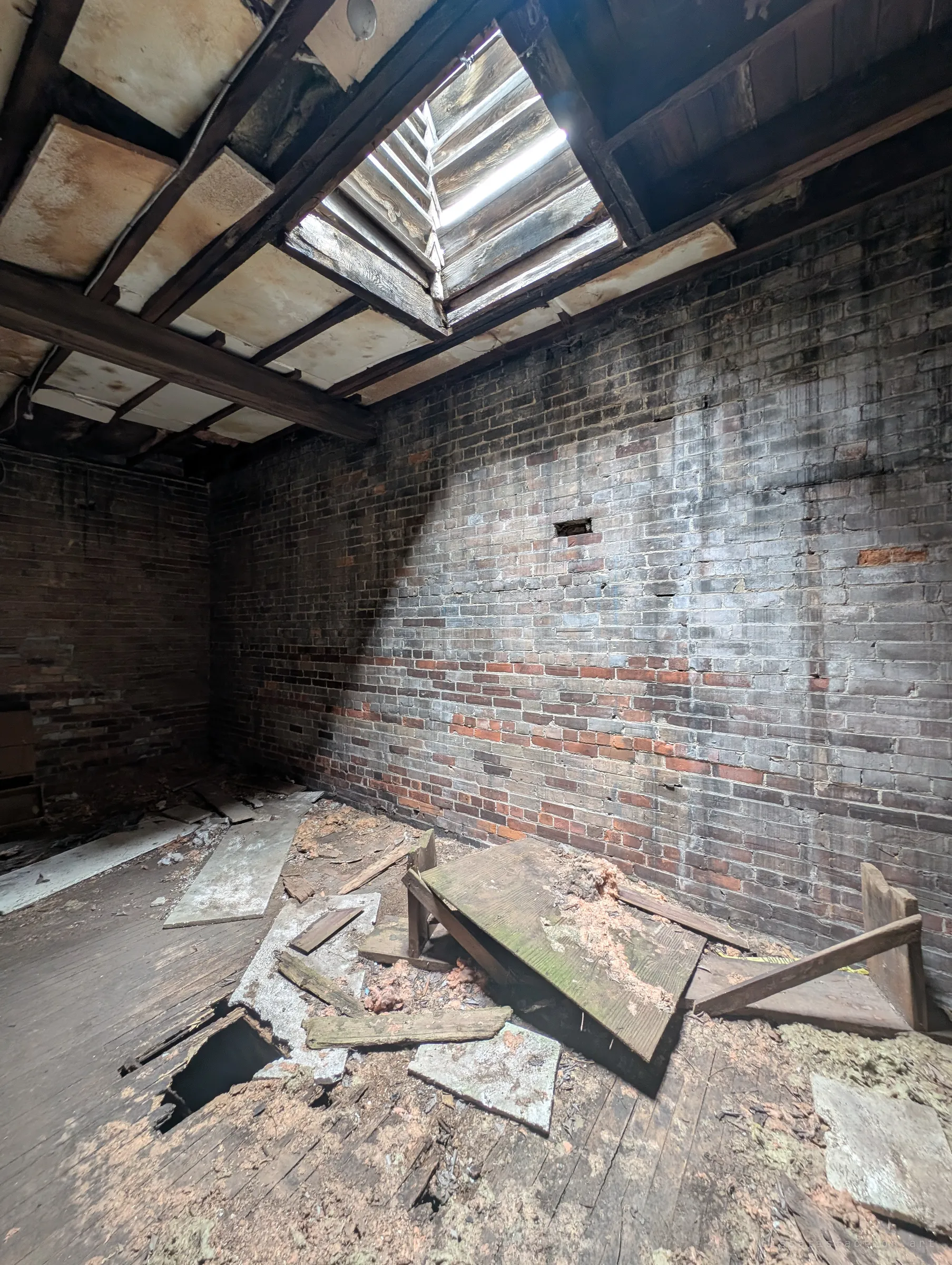 Daylight pours through a slatted roof opening onto a stained brick wall, above a rotting floor with a hole punched through it and broken boards piled around. The decay has opened the room to the sky.