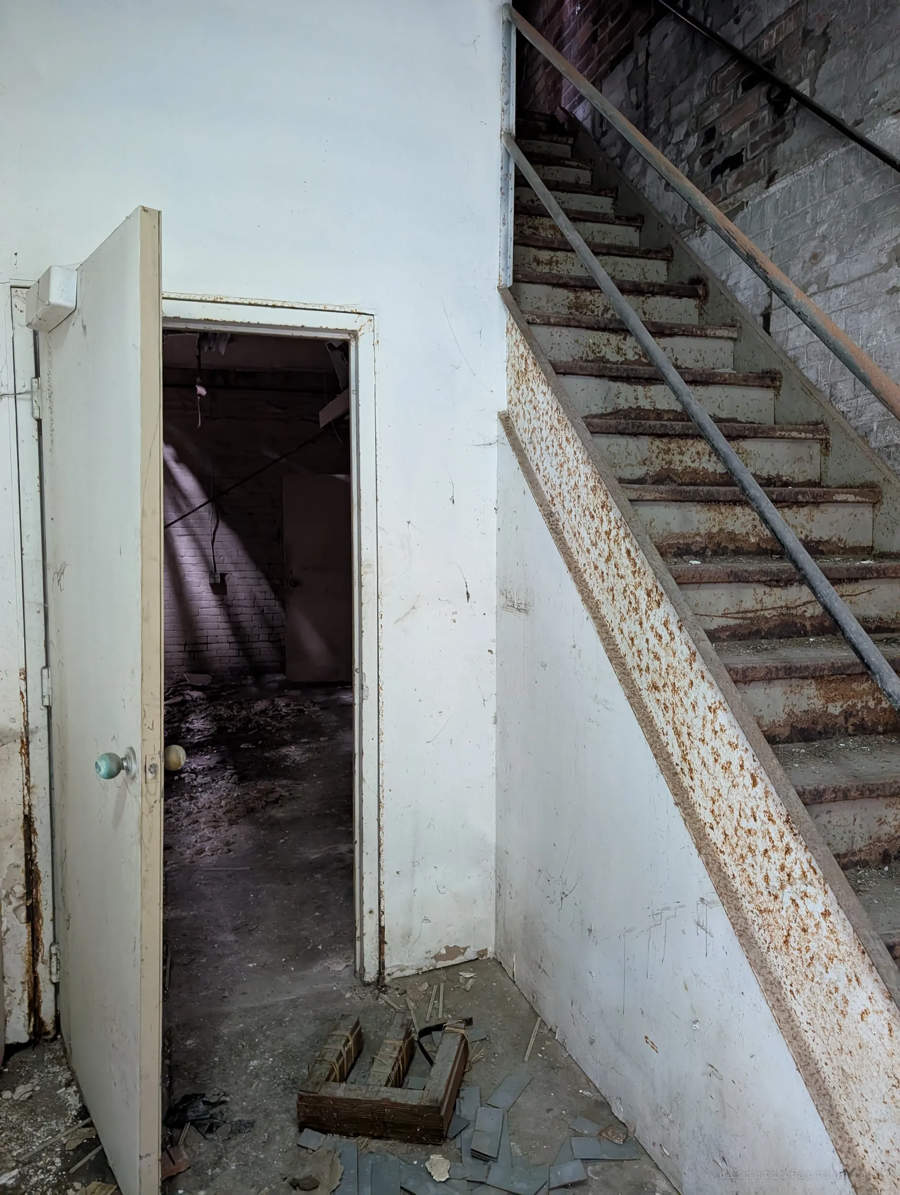 A white doorway opens onto a dim room striped with light, beside a steel staircase blistered with rust climbing along a brick wall. The rust bloom on the stringer shows years of damp.