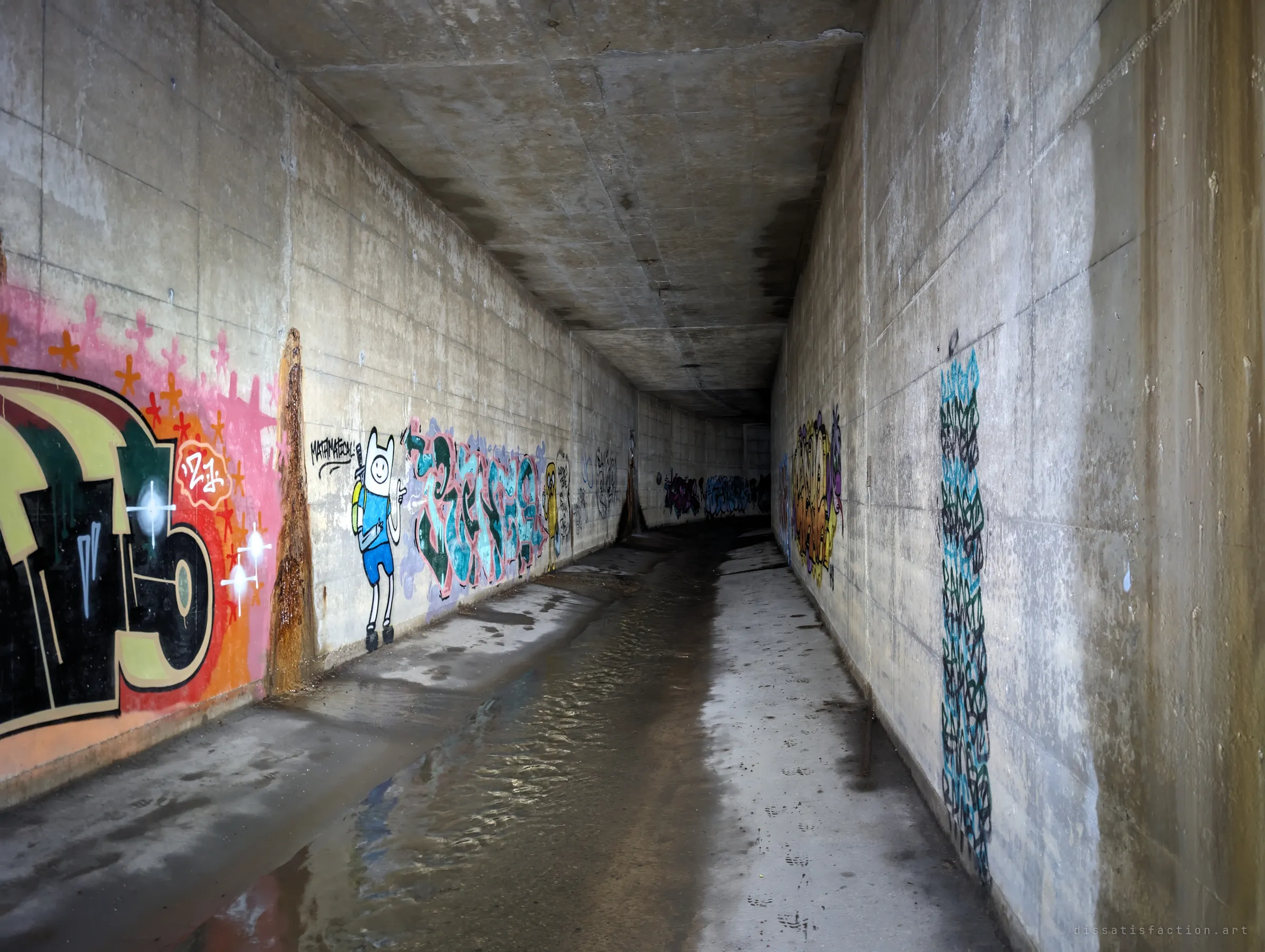 A long rectangular concrete tunnel lined with colourful graffiti, including a cartoon character, a shallow stream running down the middle into the dark.