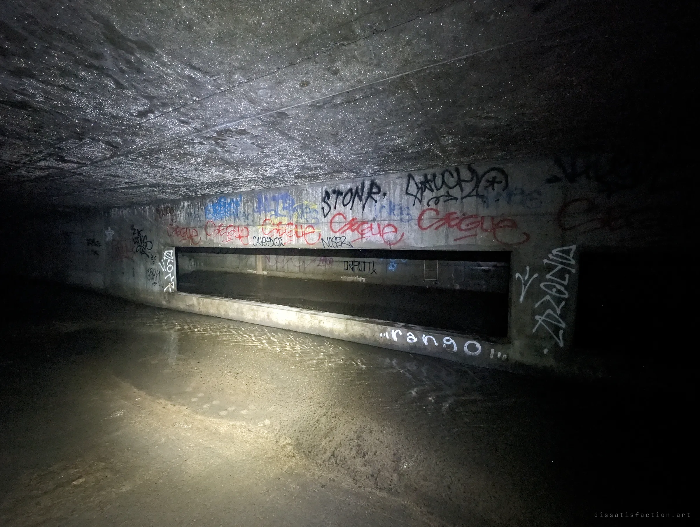 Torchlight picks out a long low opening in a tagged concrete wall, water sheeting across the floor of the chamber below a wet, glittering ceiling.