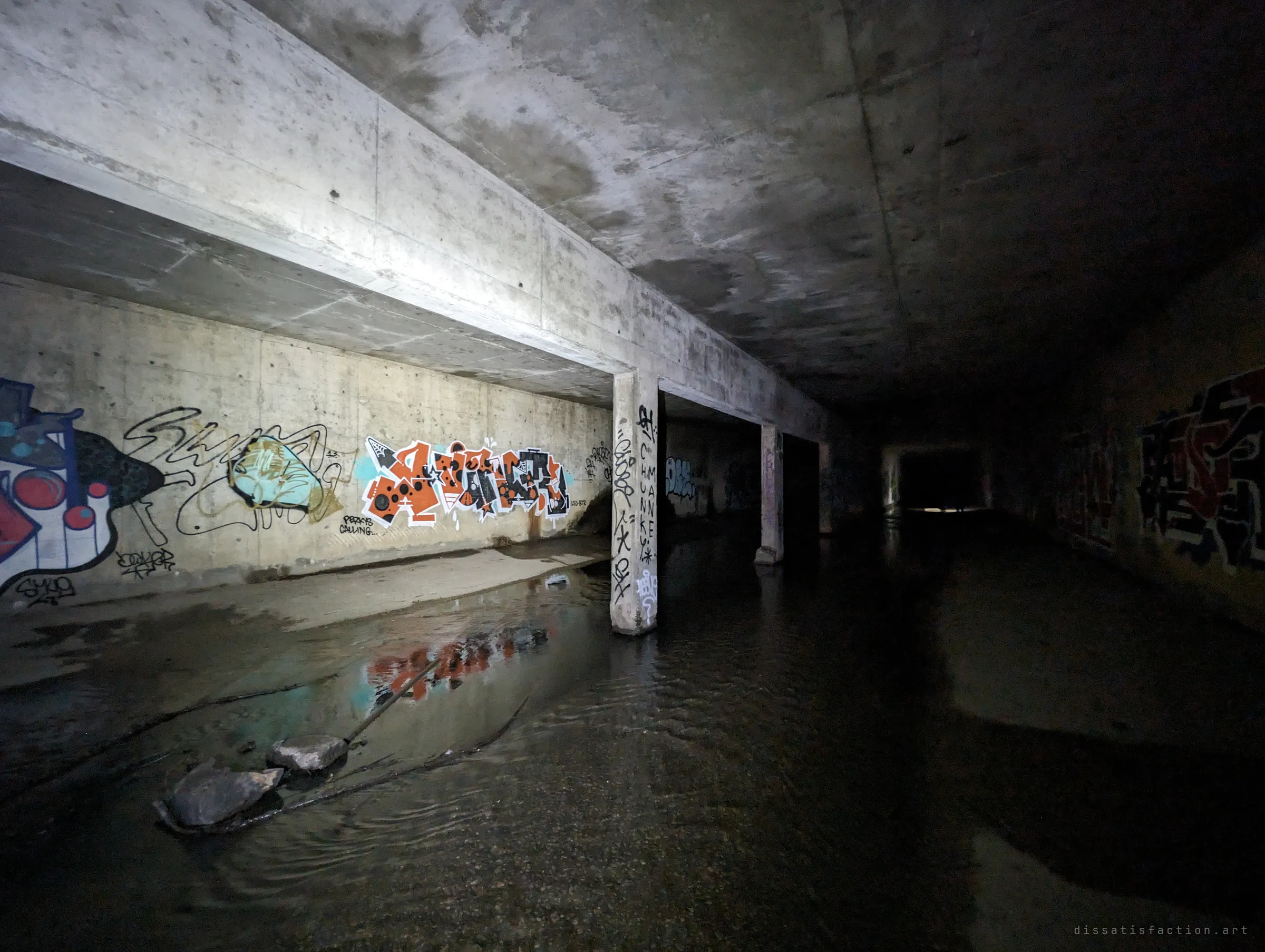 A wide underground chamber split by concrete columns and beams, graffiti pieces reflected in standing water, a faint glow at the far end.
