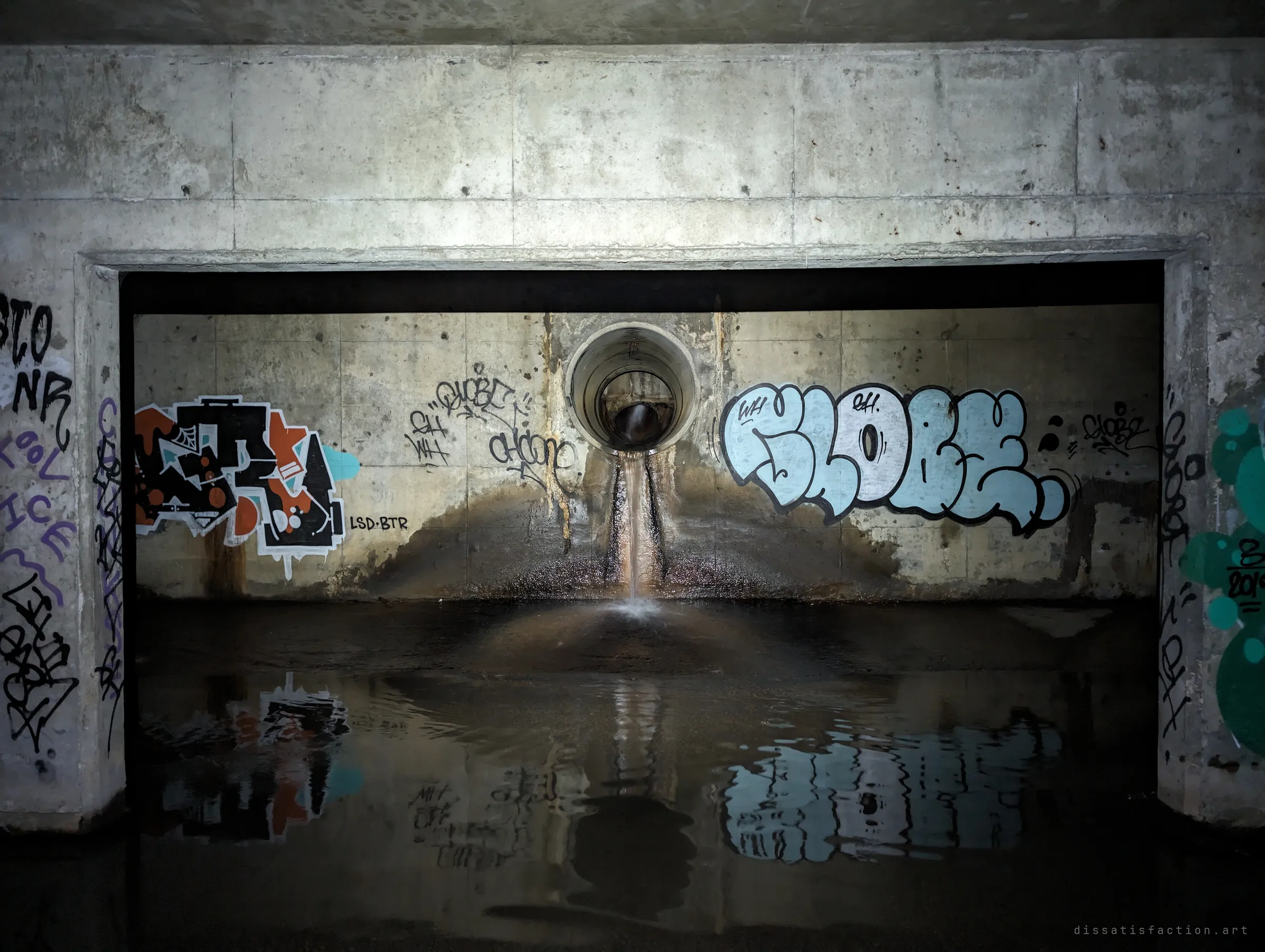 A small round pipe pours a thin stream into the chamber between two graffiti pieces, the whole wall mirrored in the still water in front.