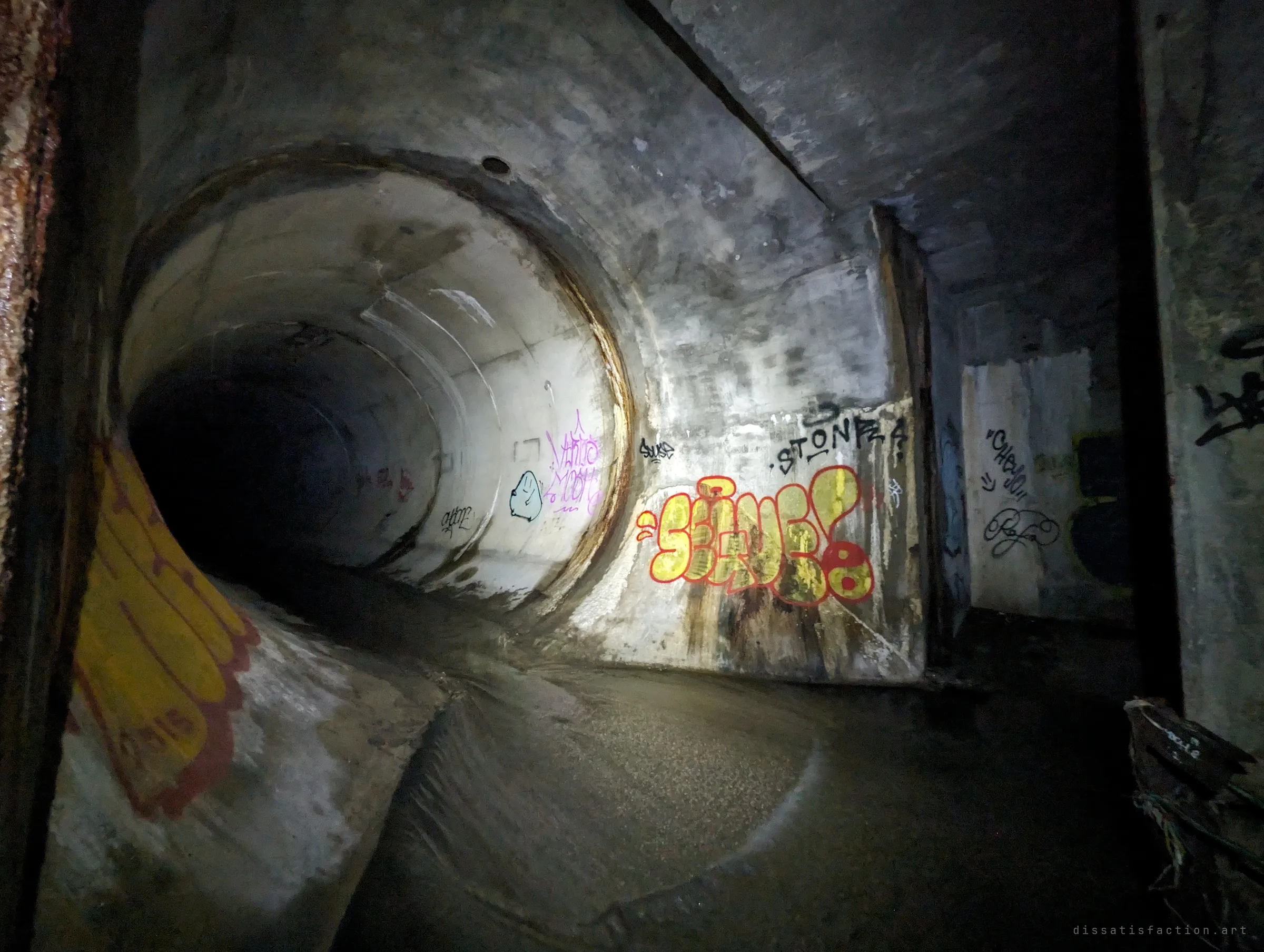 A large round concrete pipe opens out of the box chamber, its mouth ringed with rust and graffiti, water curving along the floor into the dark.