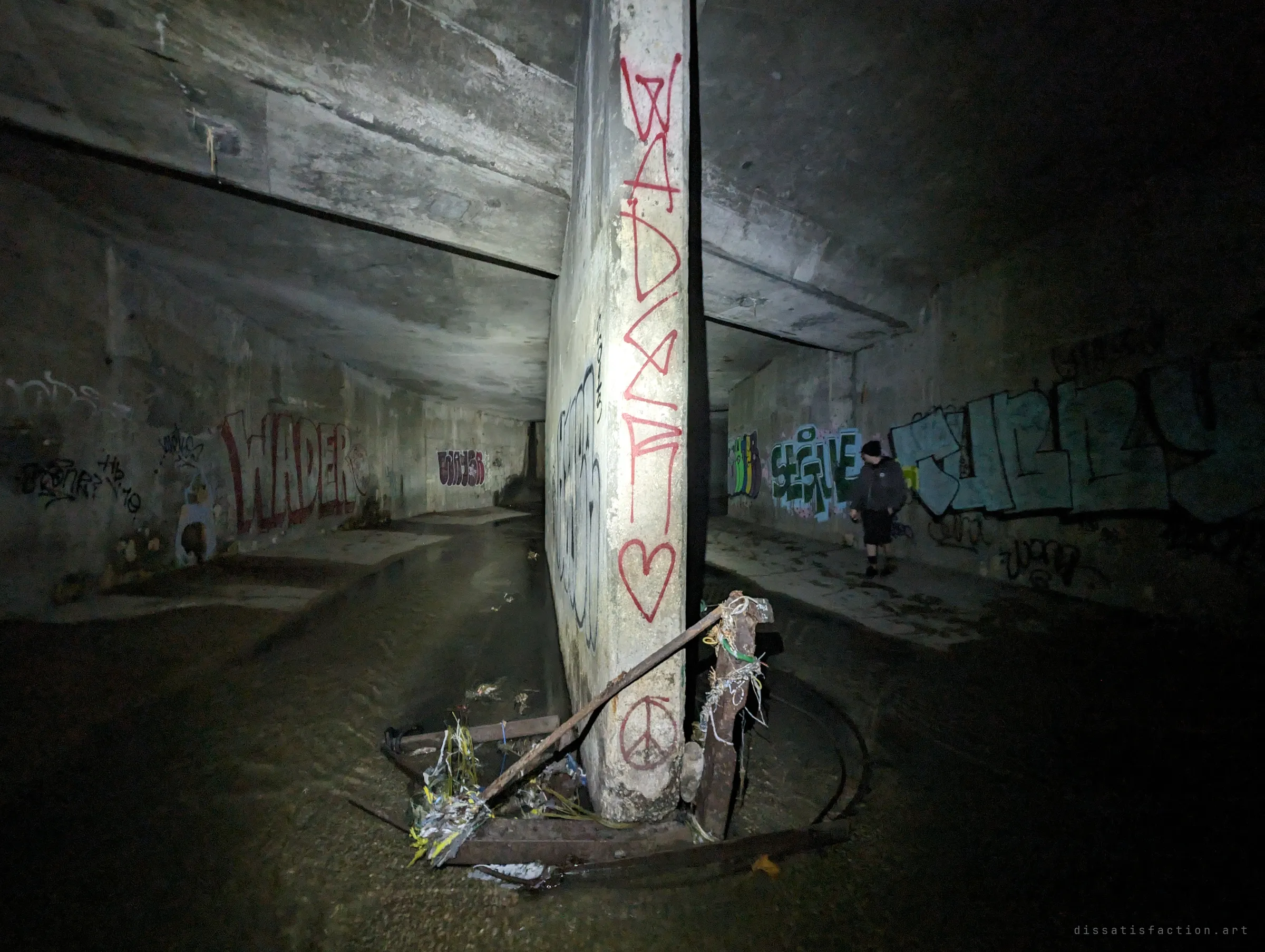 A graffiti-covered central column splits the chamber into two channels, wrecked metal debris tangled at its base; an explorer in a dark hoodie walks along the right-hand wall for scale.
