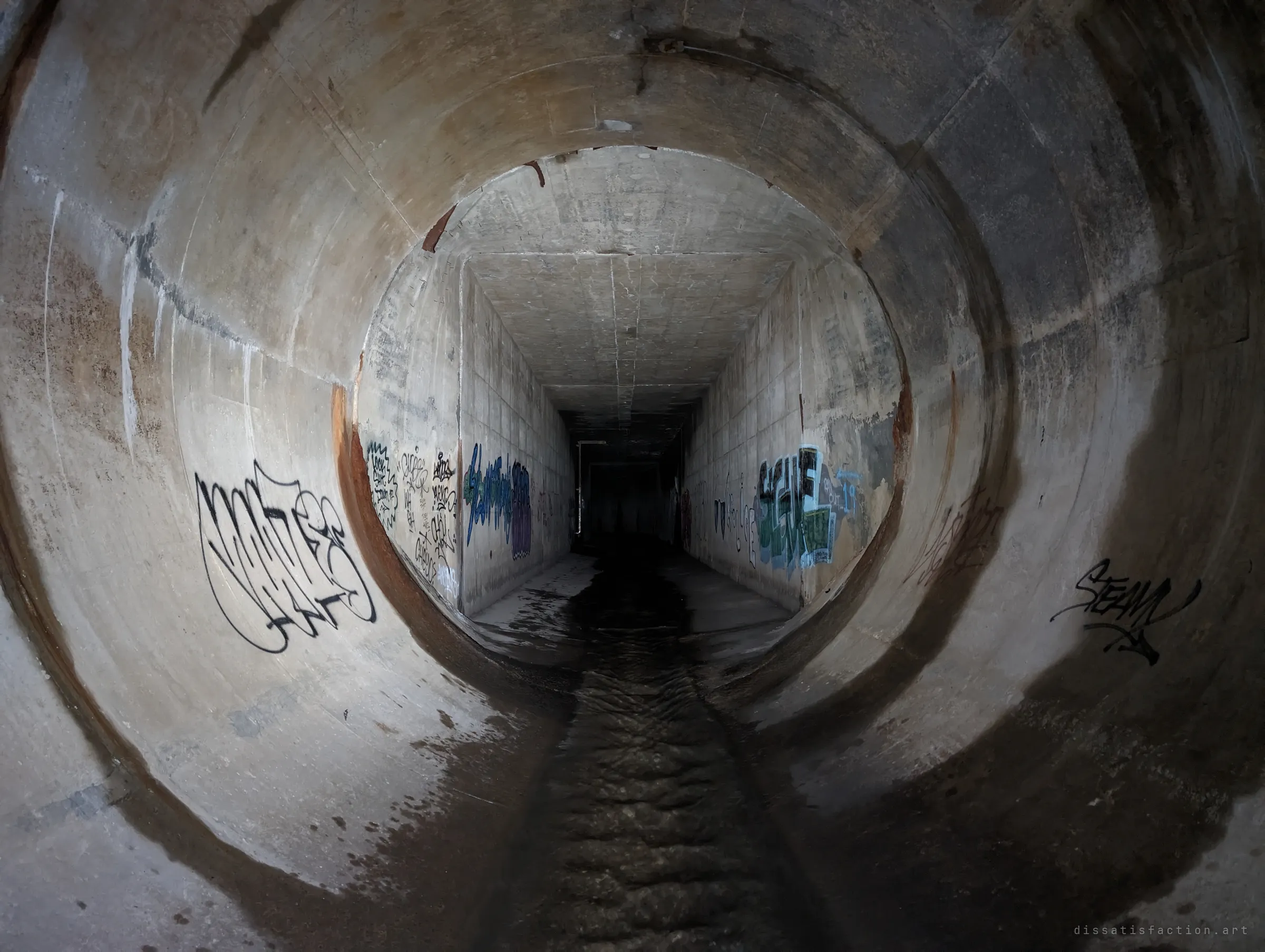 A round concrete pipe opens into a square box culvert covered in graffiti tags, a thin stream of water running down the middle. The change of shape marks where two eras of drain construction meet.