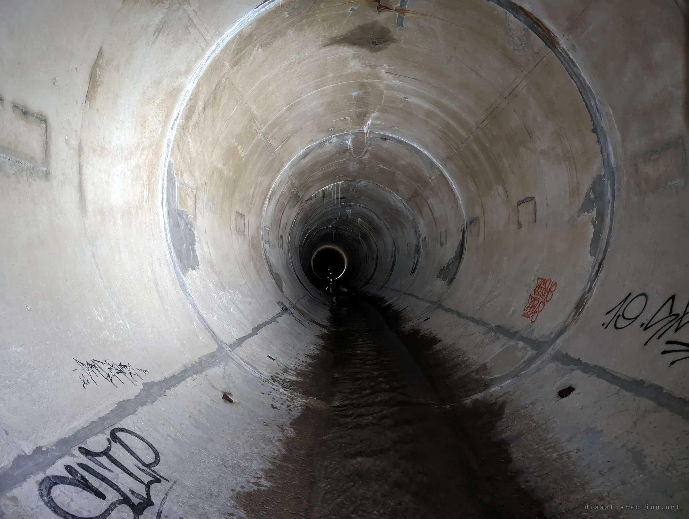 A long round concrete pipe recedes in stepped rings toward a tiny dark opening far ahead, with water trickling along the floor and a few tags on the walls. The repeating rings show how deep the system runs.
