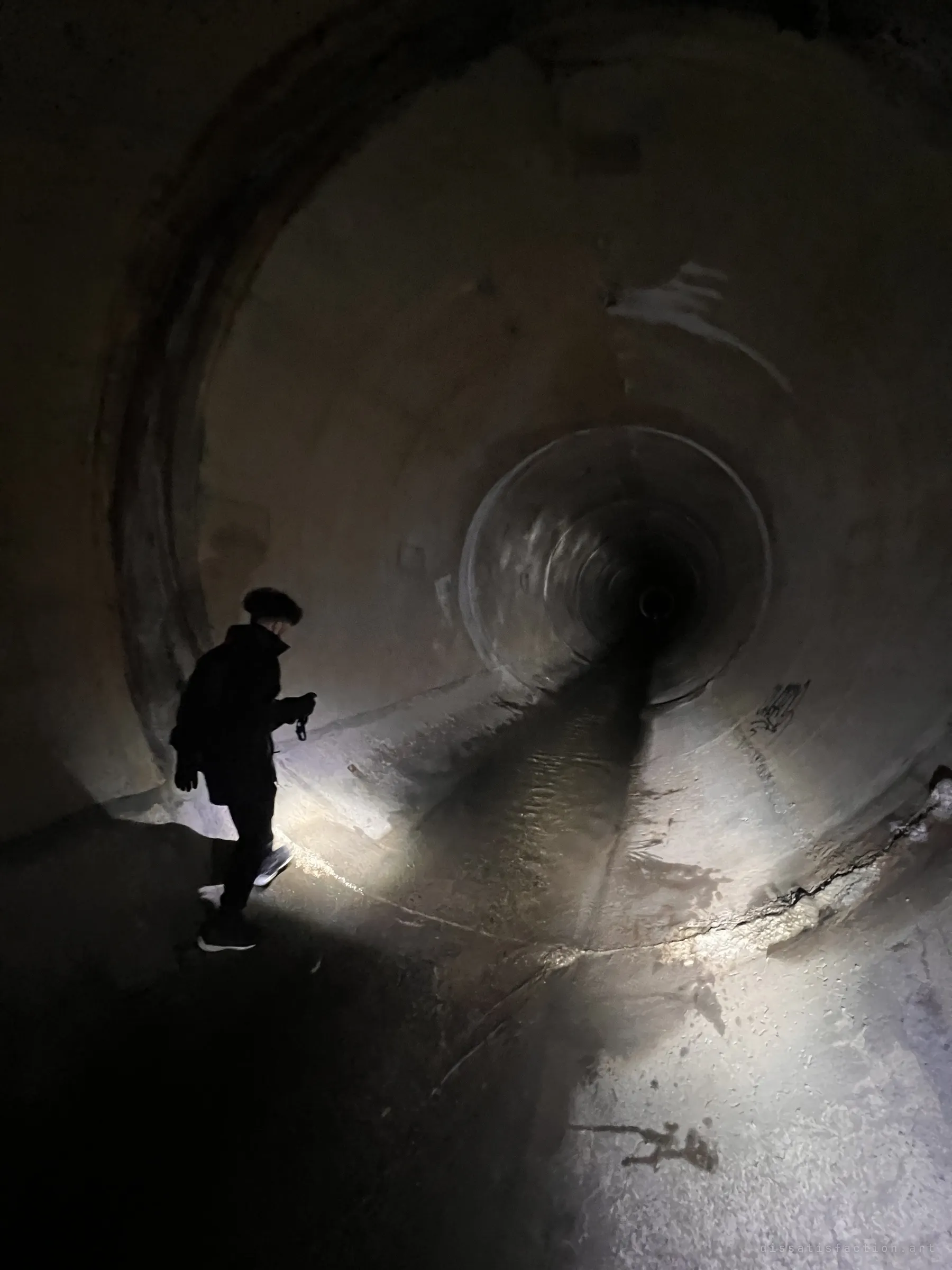 An explorer in dark clothes stands at the edge of a wide pipe, torch in hand, looking toward a smaller pipe that feeds in from the far wall. The torch beam lights the wet, cracked floor.