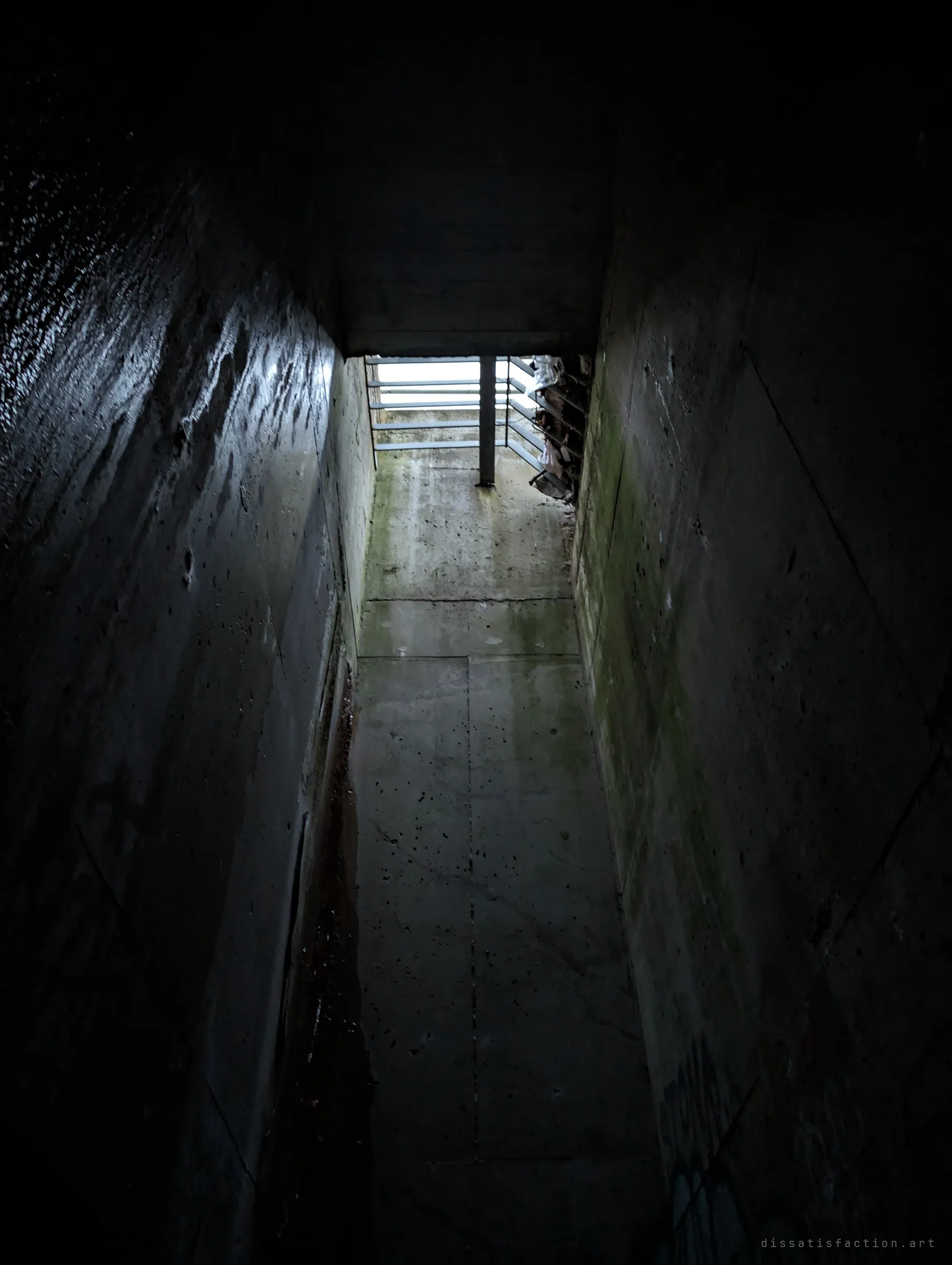 A narrow concrete channel with moss-streaked walls leads up toward a barred opening and daylight. It is the way in or out of the drain.