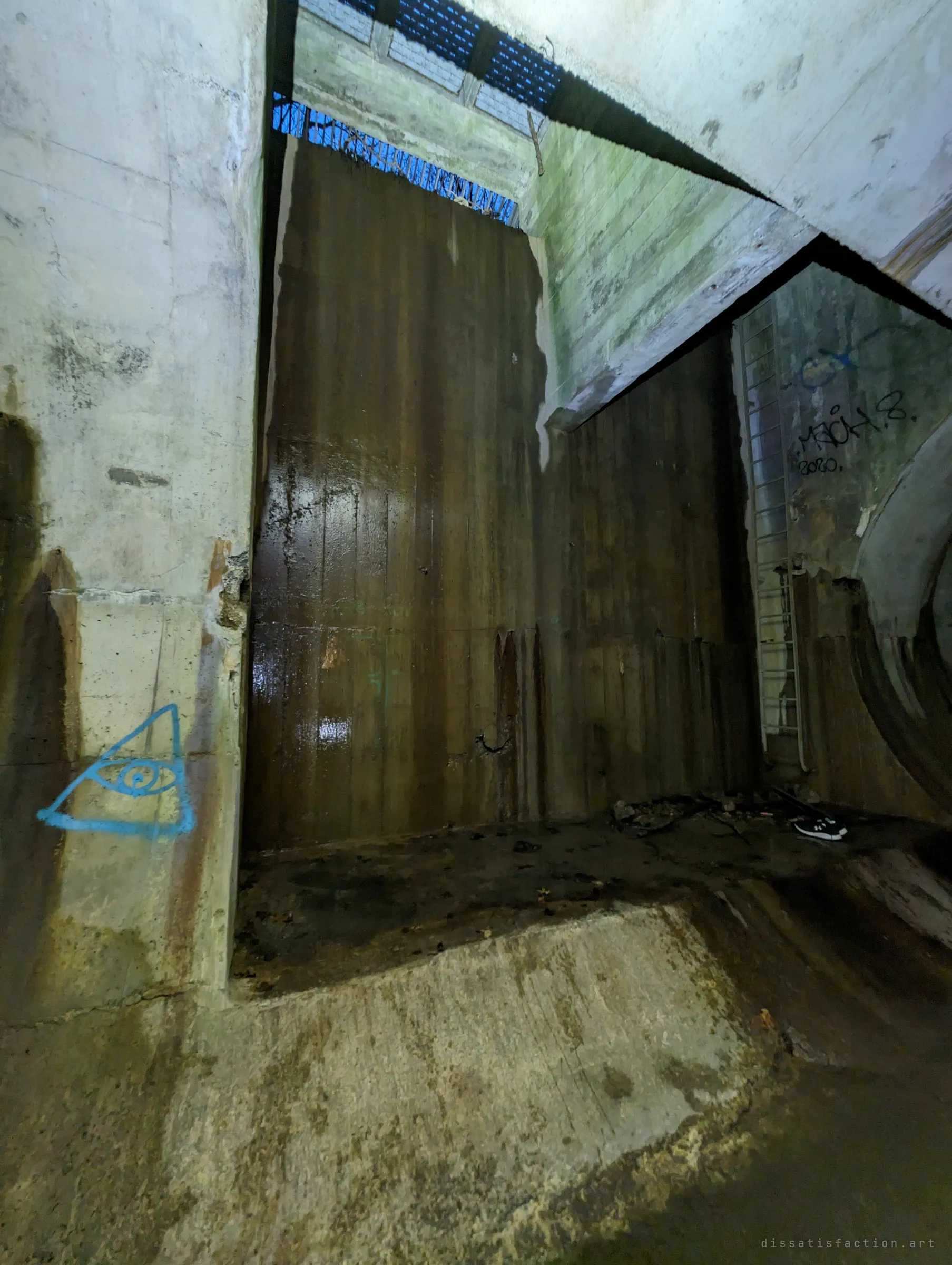 A tall chamber with a rust-stained steel wall, a grated opening high above letting in blue light, a ladder on the right and a painted eye-in-a-triangle on a concrete pillar. Debris and a lost shoe lie on the floor.