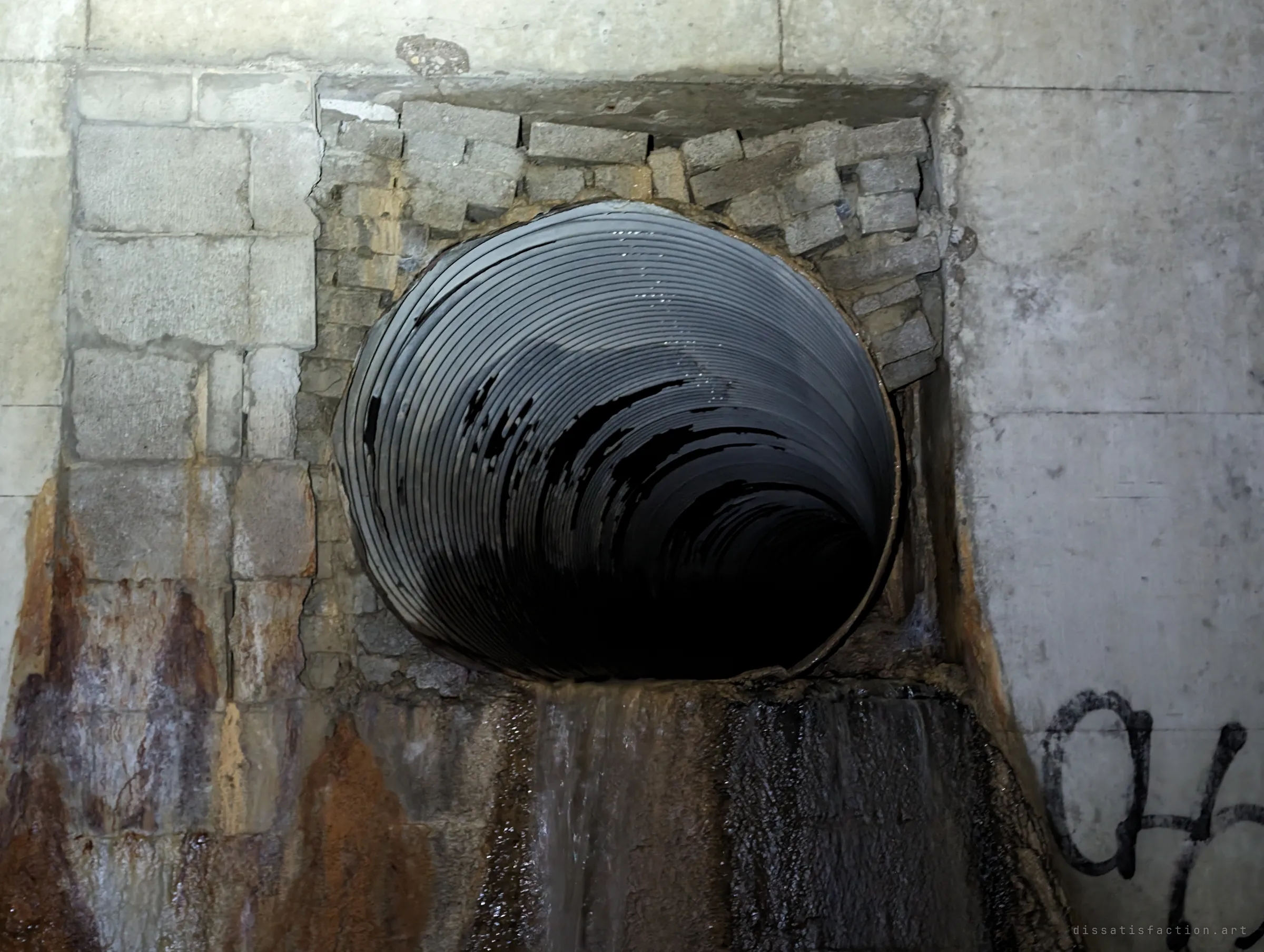 A corrugated metal pipe pokes through a rough patch of stacked concrete blocks and spills a thin stream of water down a rust-stained wall. The makeshift fit shows the drain being altered over time.