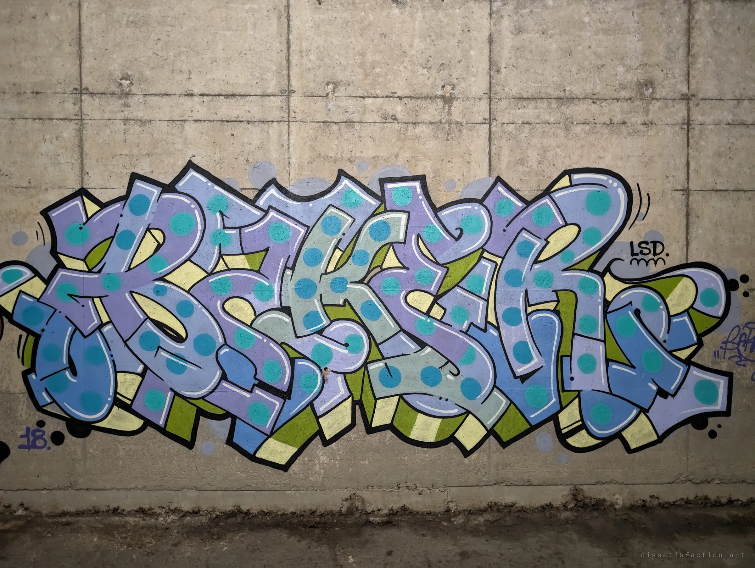 A large wildstyle piece in lavender, blue and teal polka-dot letters fills a board-marked concrete wall. The tunnel walls work as a sheltered gallery for local writers.