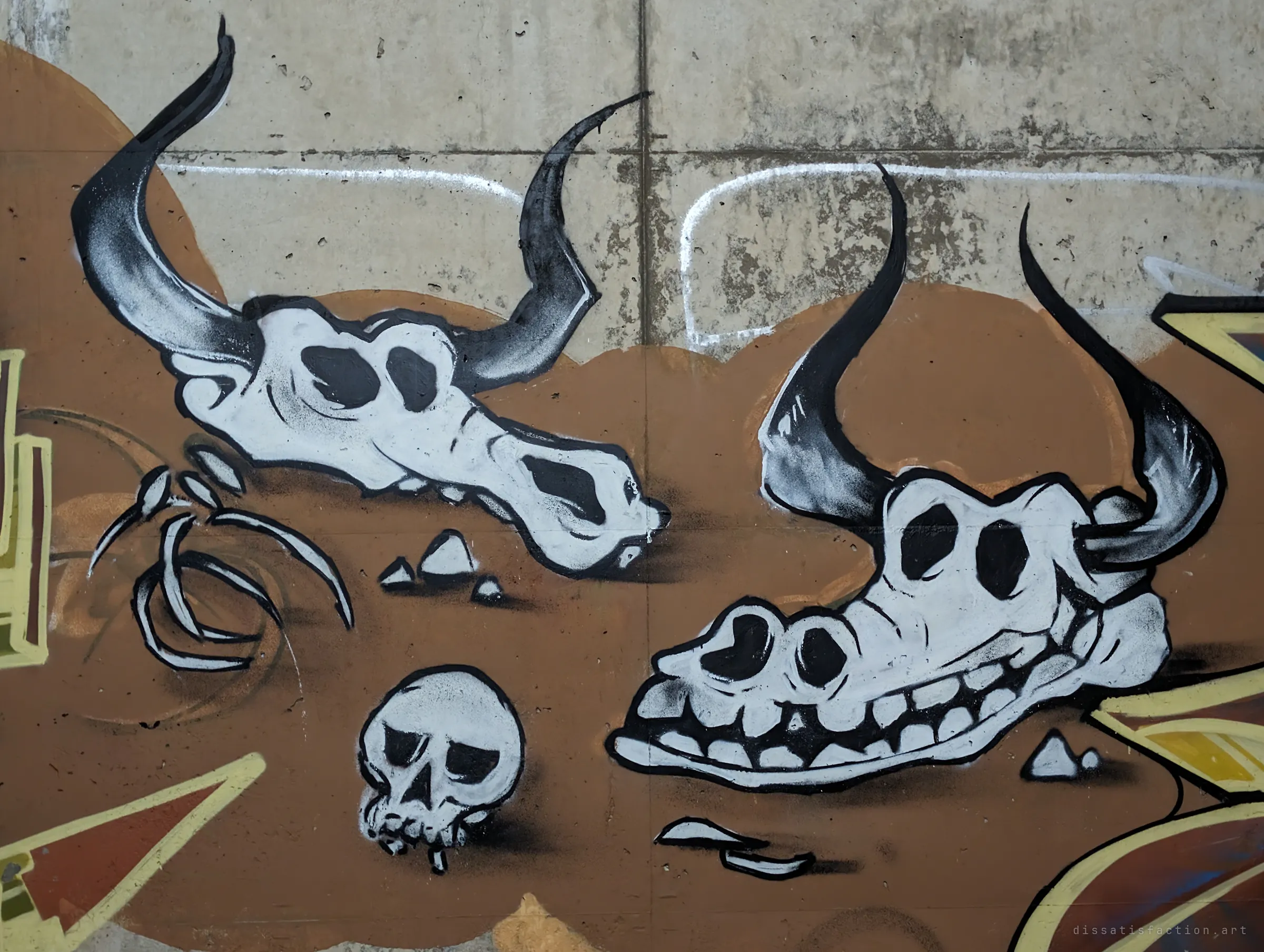 Painted cartoon cattle skulls and a small human skull lie on a brown desert backdrop across the concrete. A darkly playful character piece among the letter walls.