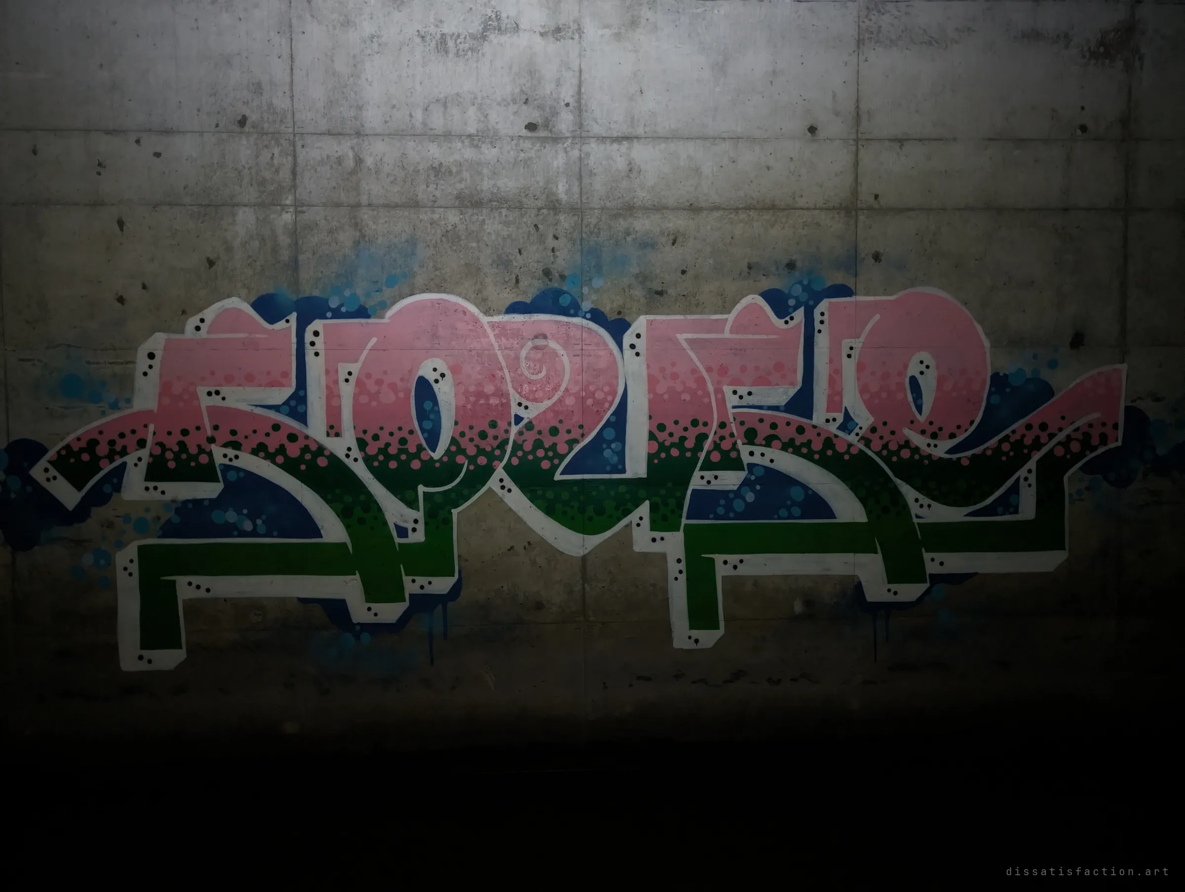 A pink-to-green dotted letter piece glows under torchlight on the formwork concrete while the floor falls away into black. Only the lit patch of wall shows how dark the passage is.
