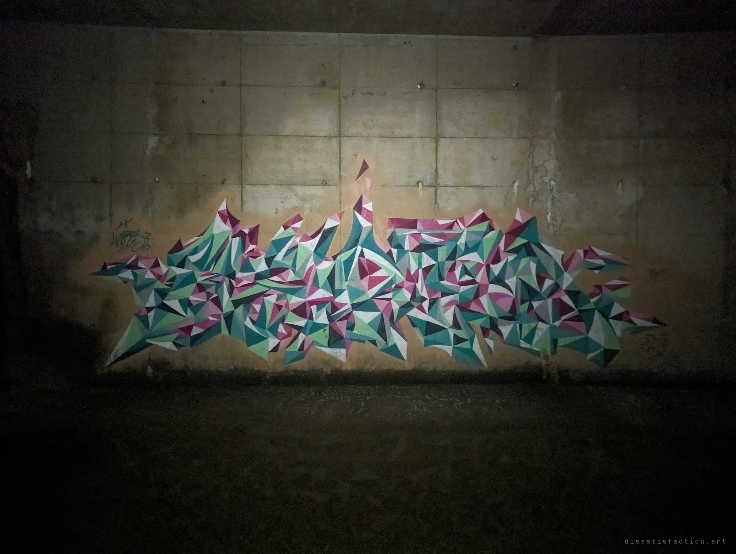A faceted piece of teal, mint and magenta triangles emerges from darkness on the tunnel wall. The torch beam picks it out while the floor stays black.