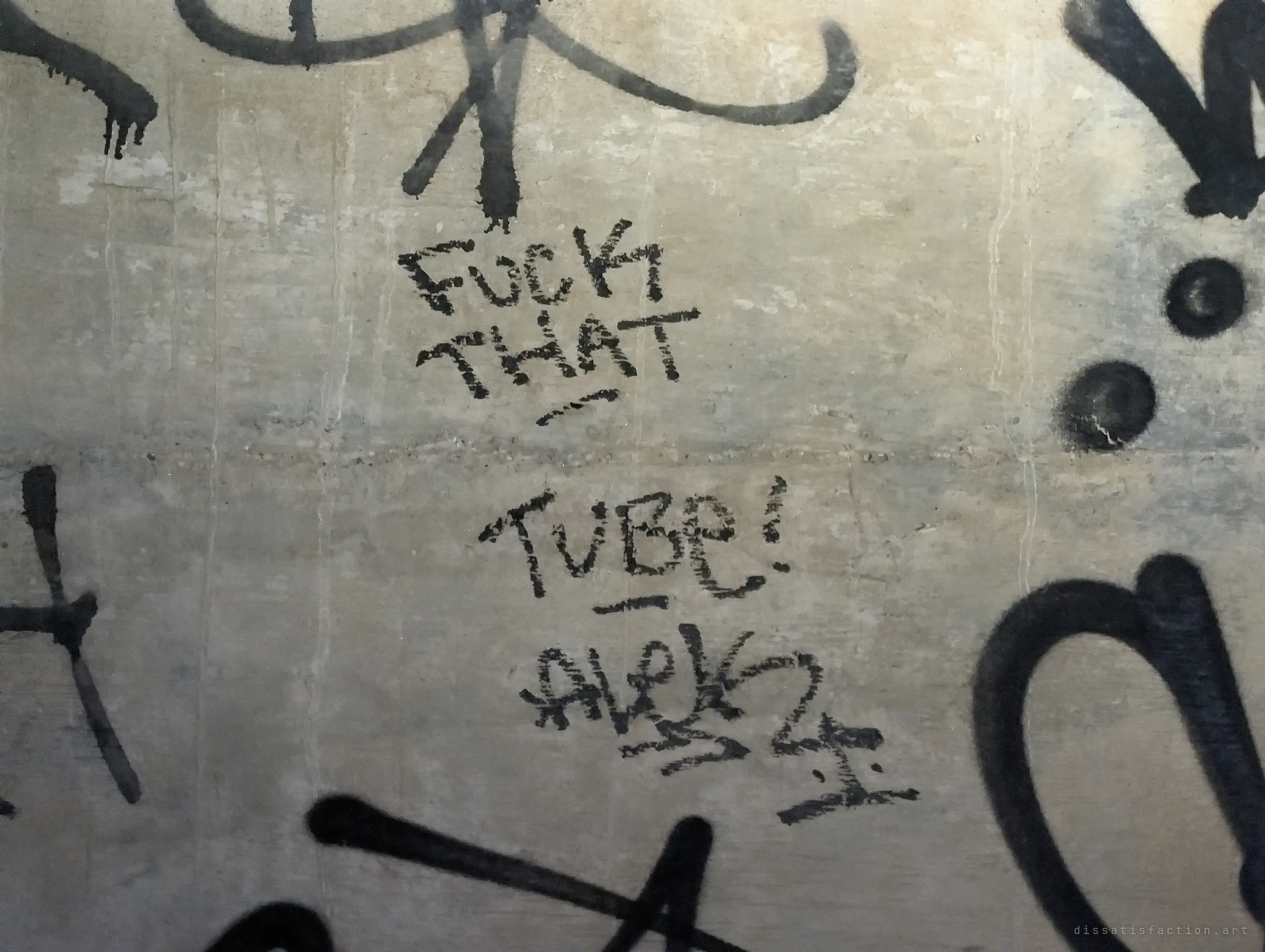 Crude marker scrawl mocking the tunnel sits among heavy black tags on the concrete. A small, rude note from someone who passed through.