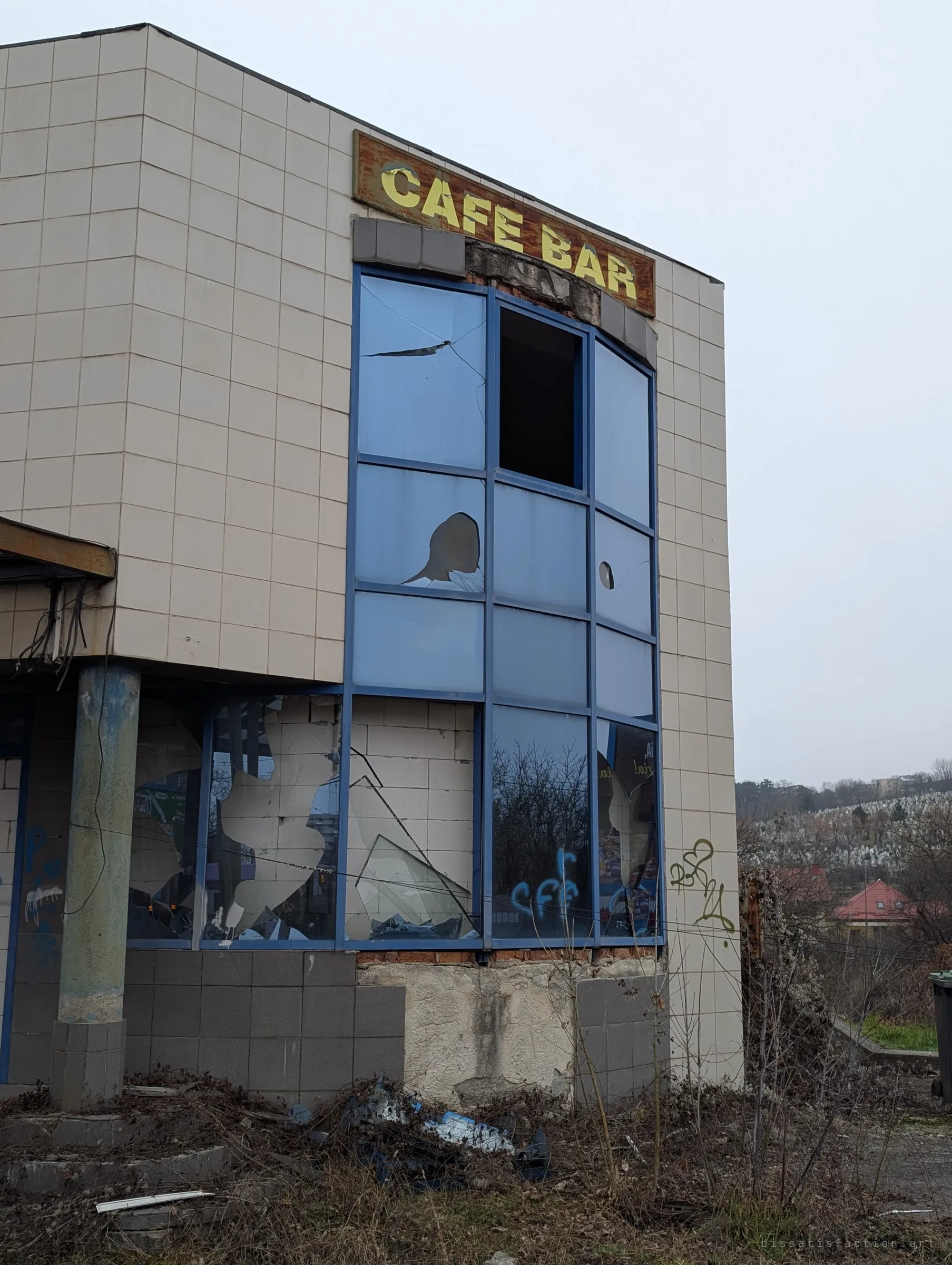 The tiled corner of an abandoned roadside cafe bar, its blue curtain-wall glazing smashed and a rusty sign still hanging above. Shows how far the building has decayed behind its once-modern facade.