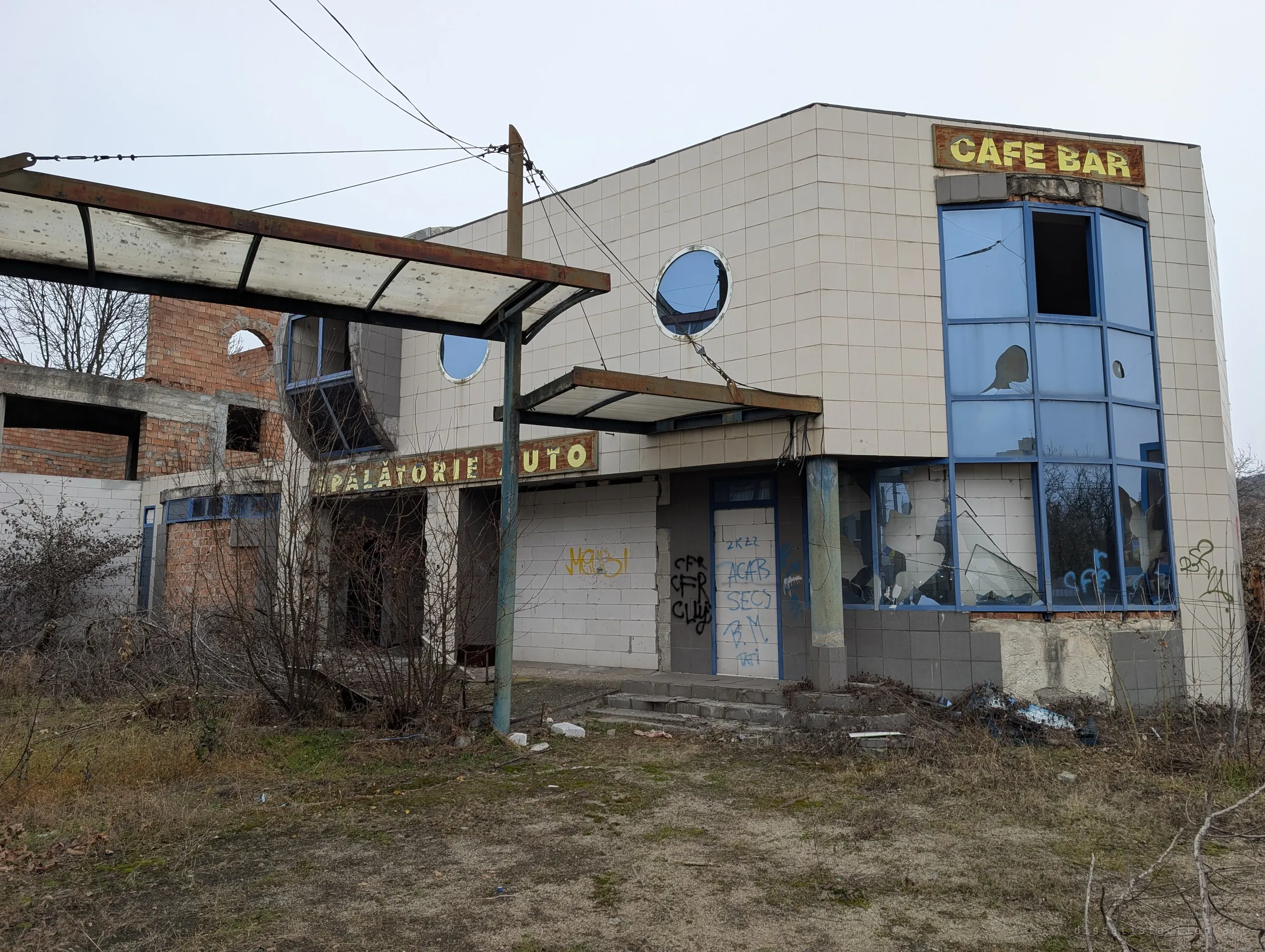 Wide view of the abandoned car wash and cafe complex, with a rusted canopy, porthole windows and an unfinished brick upper floor behind overgrown scrub. Sets the scene for the whole site.
