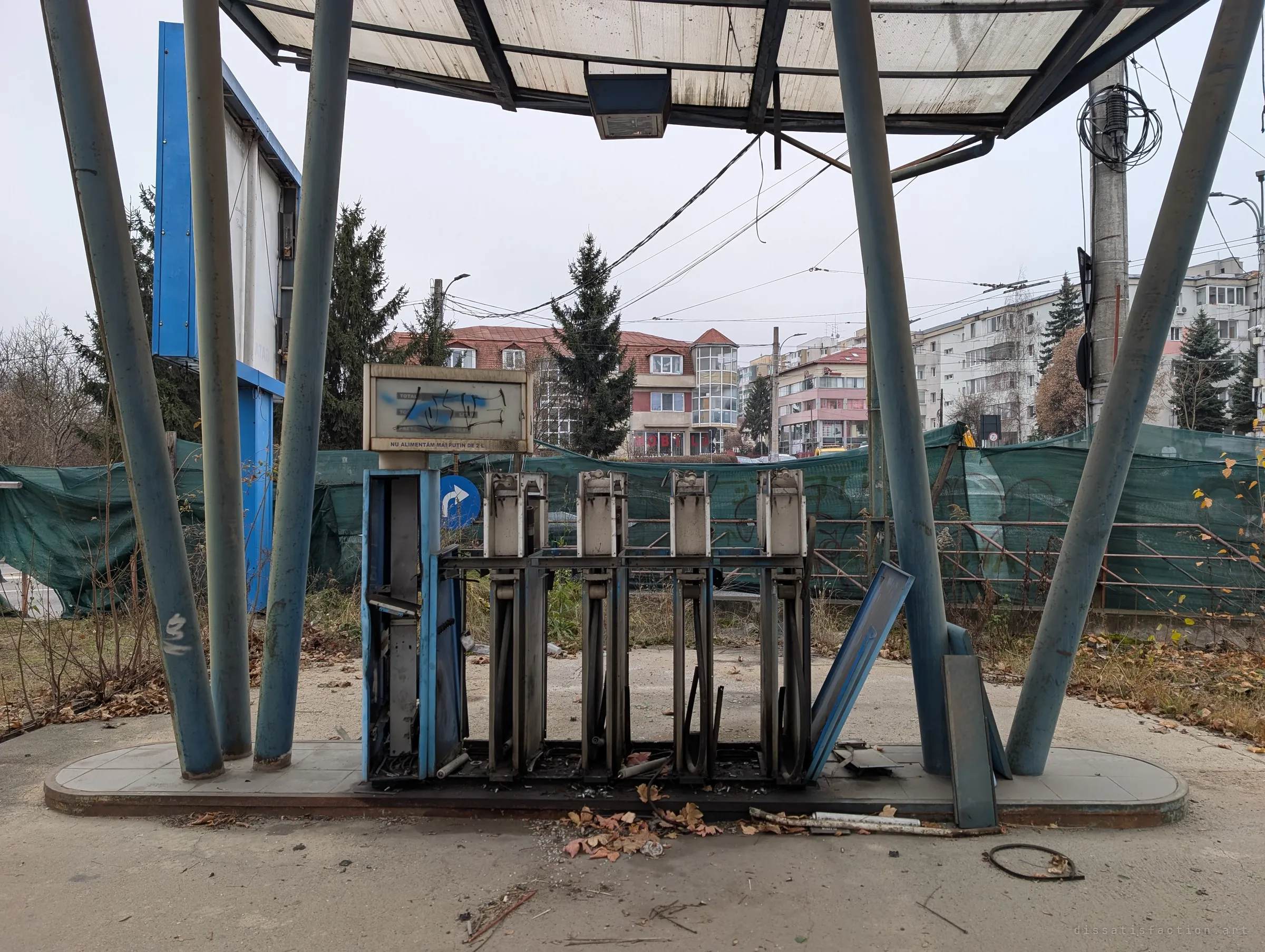 A gutted fuel pump island under a sagging canopy on slanted blue pillars, the dispensers stripped to their frames. The empty forecourt shows the station's abandonment against a busy street beyond.