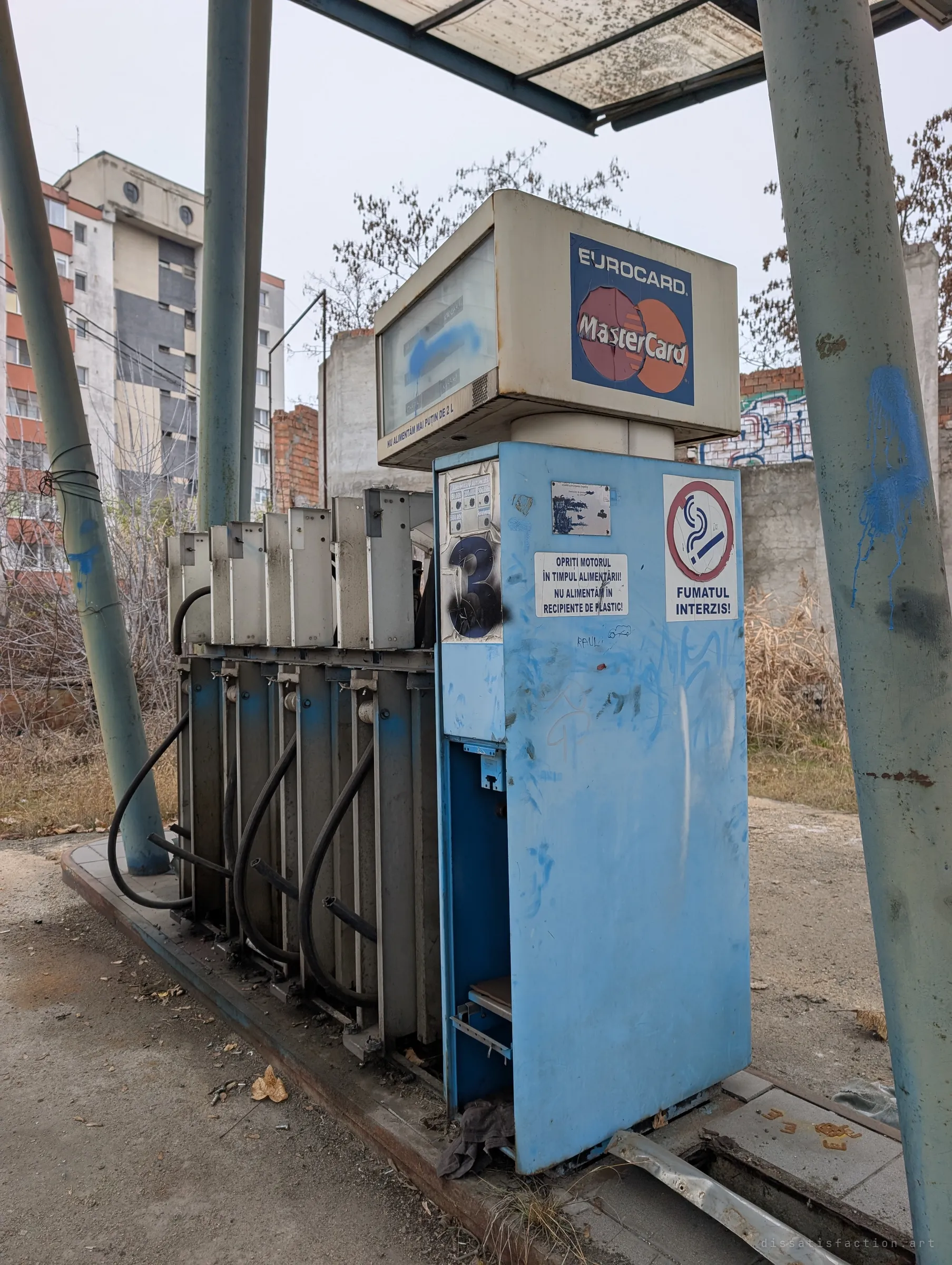 An old blue fuel dispenser with a faded card-payment sign and no-smoking sticker, its hose bays empty beside it. A close look at the station's last working equipment.