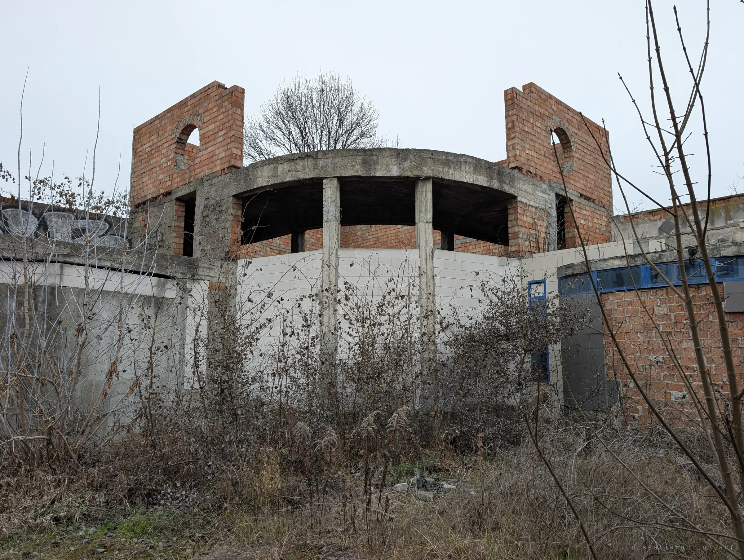 A half-built rotunda of brick and concrete columns with arched wall fragments, rising out of bare winter scrub. The abandoned construction looks almost like a ruin.