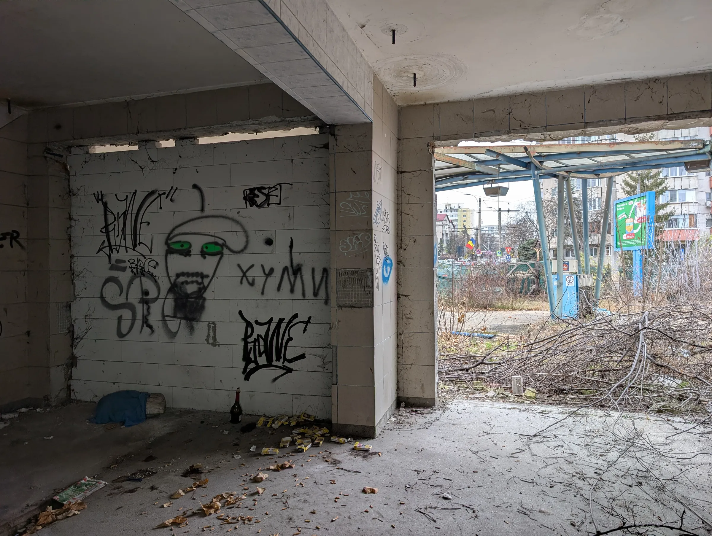 Inside the ground floor, a tiled wall covered in graffiti with a painted green-eyed face, cigarette packs and a bottle on the floor. The open doorway frames the old pump canopy outside.