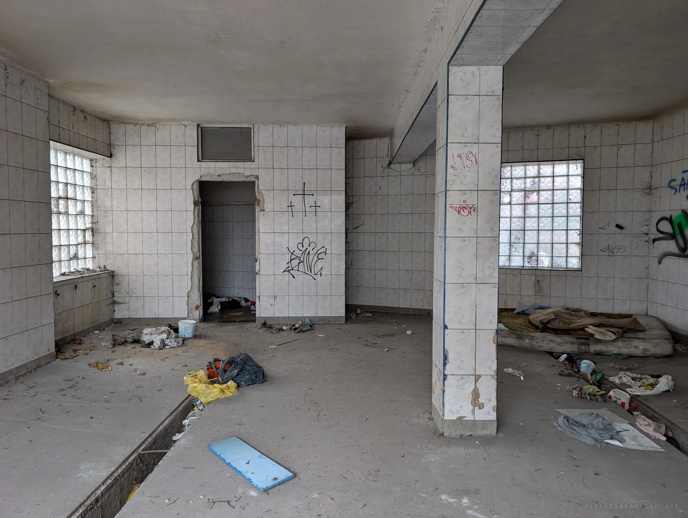 A tiled former service room with glass-block windows, an open inspection pit and a mattress with bedding among scattered rubbish. Signs that people have been sleeping here.