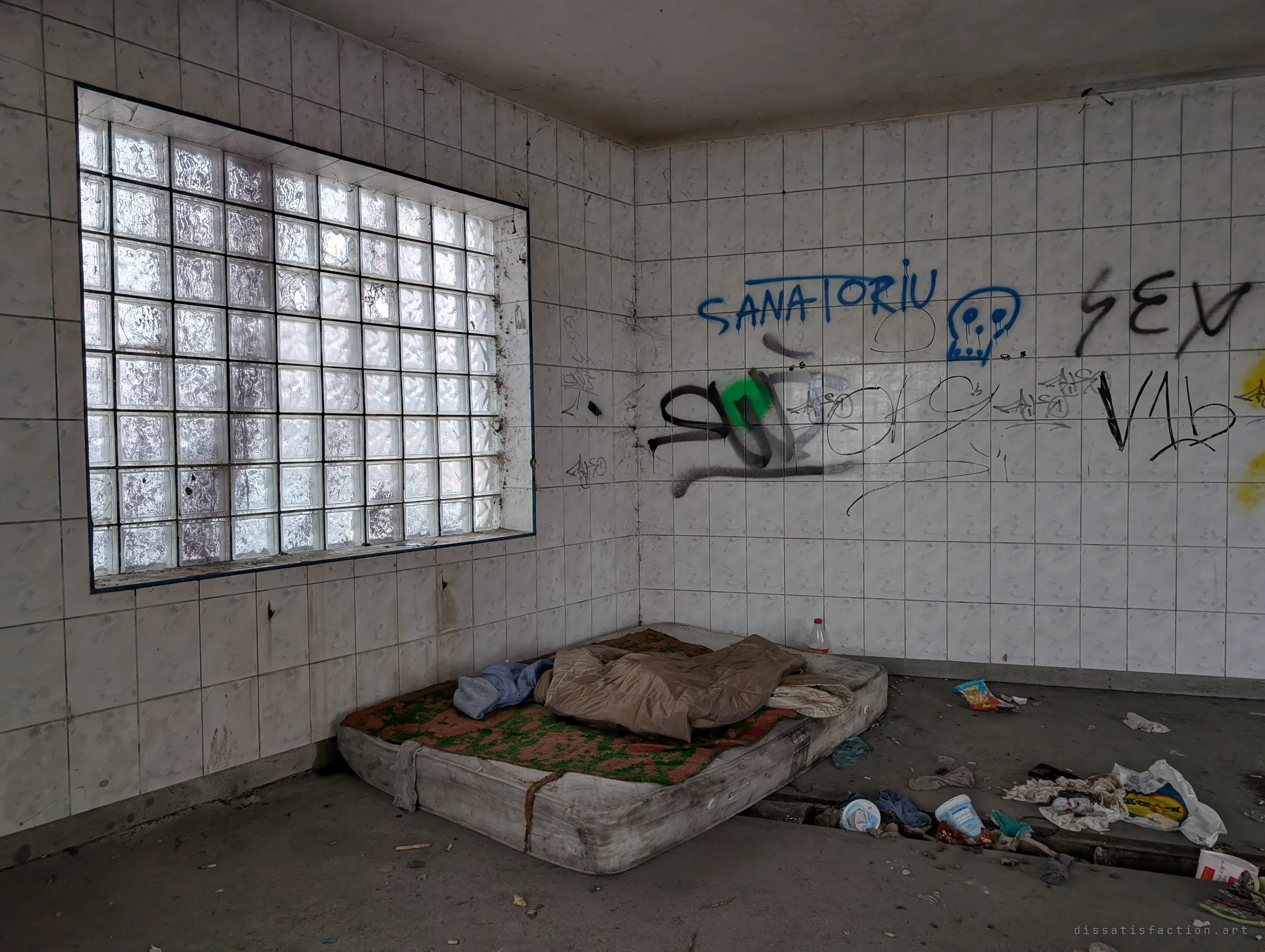 A mattress and sleeping bag in the corner of a tiled room under a large glass-block window, the walls tagged with graffiti. A quiet, lived-in corner of the derelict building.