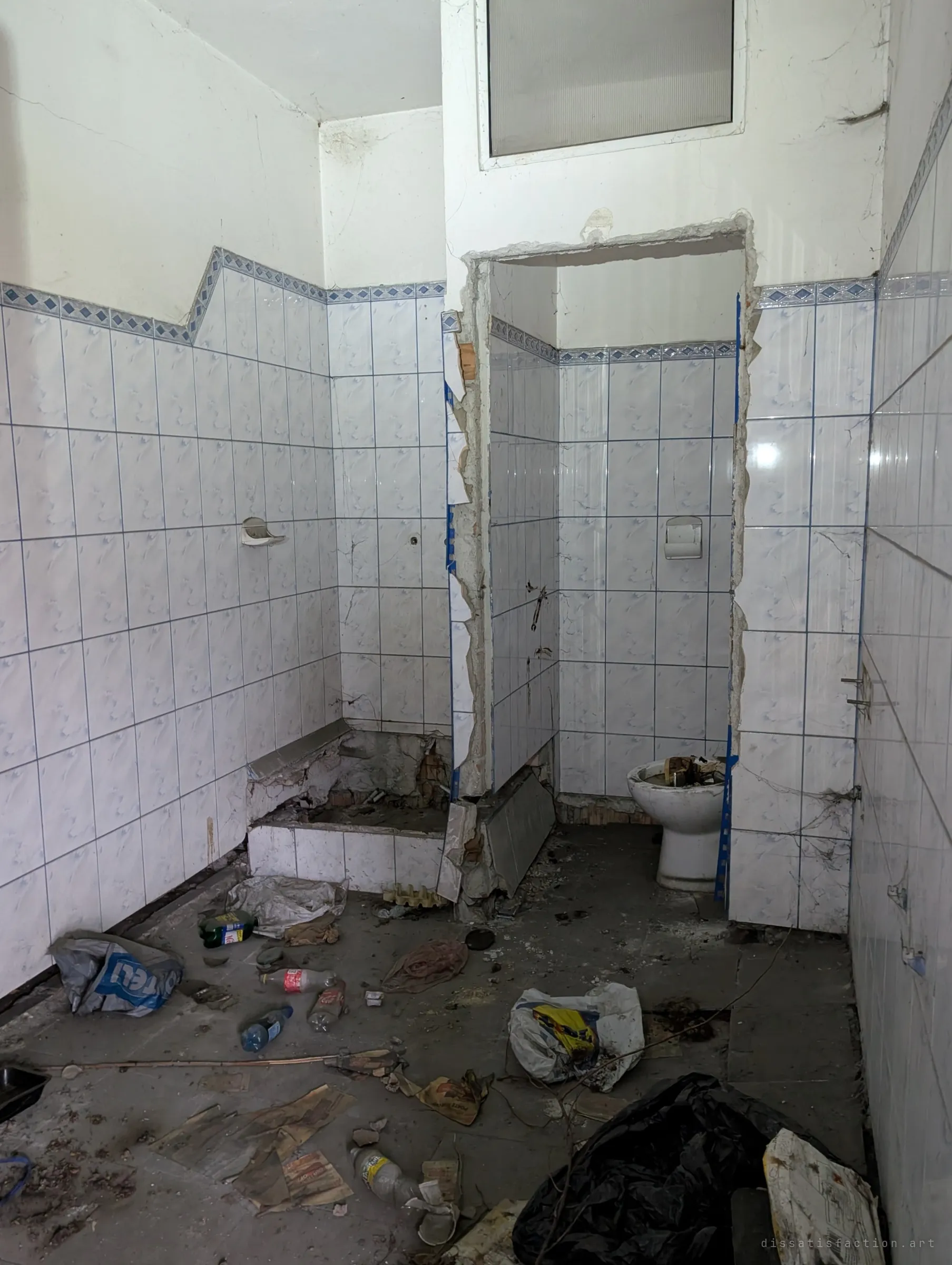 A gutted tiled washroom with its stall doors torn out, the floor strewn with bottles, bags and wet paper. The ripped-out door frames show how thoroughly the building has been stripped.
