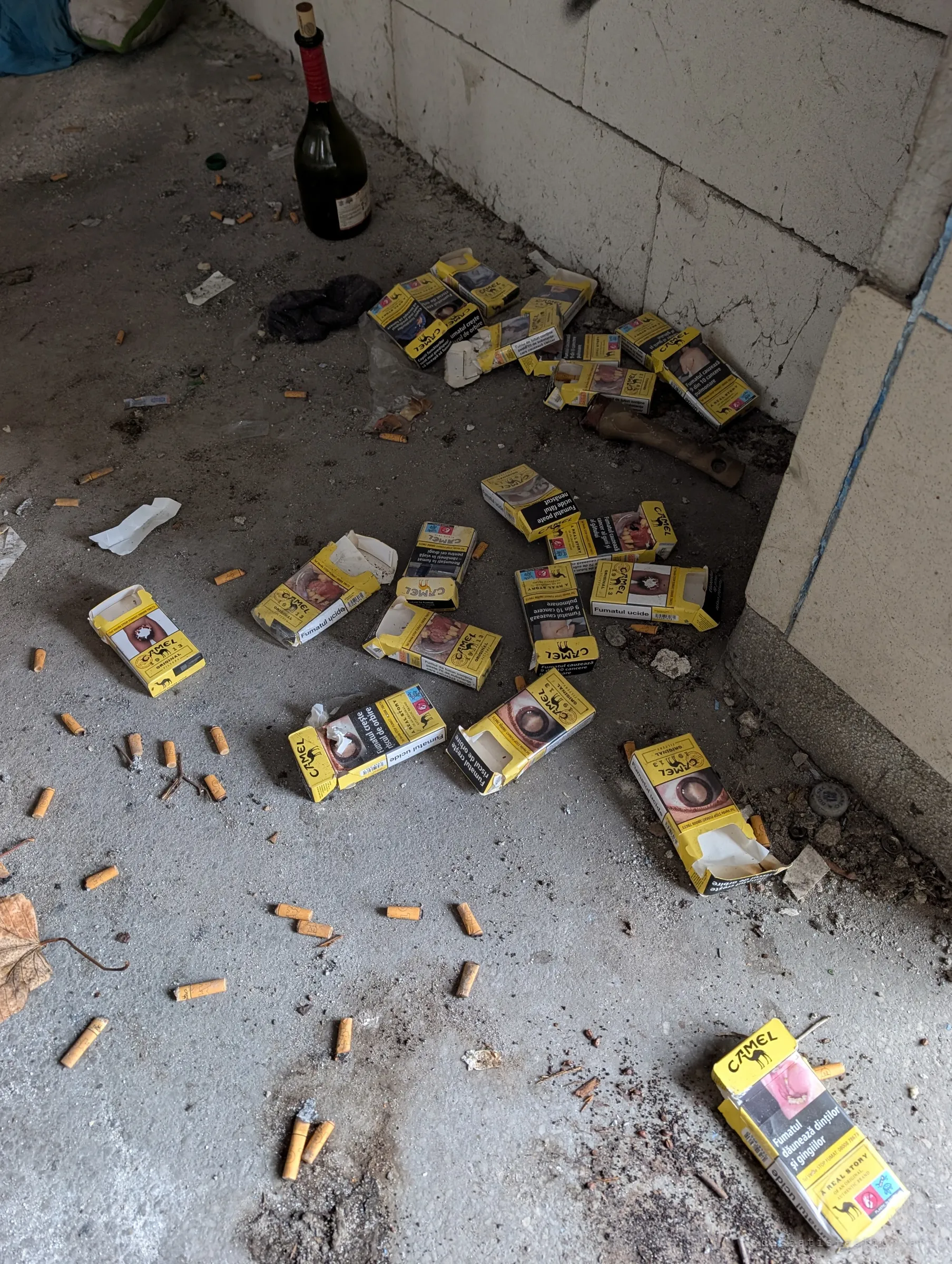 Dozens of empty cigarette packs and butts scattered across a concrete floor beside a wine bottle. It is the leftover of someone who spent long nights here.