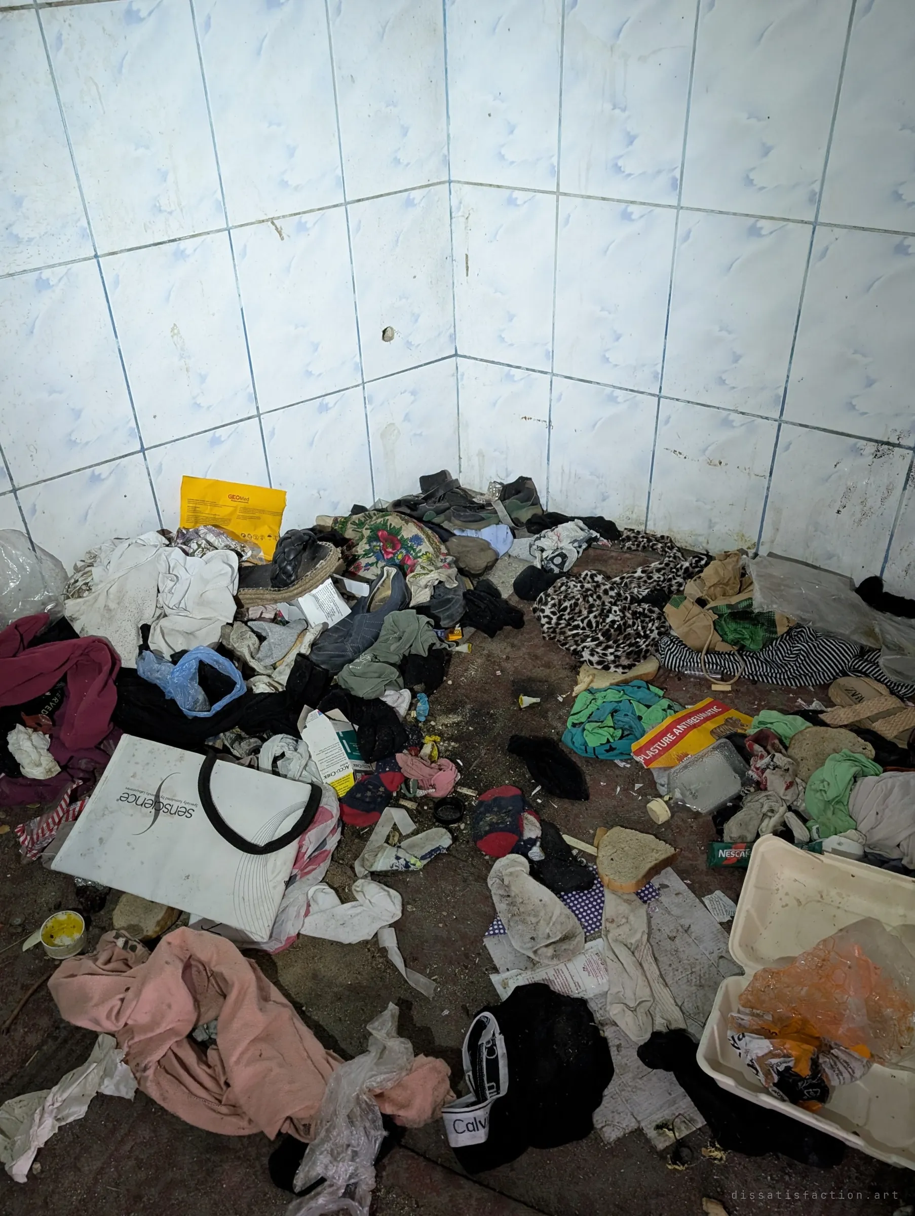 A tiled corner heaped with clothes, shoes, medicine packaging and a food container. Someone has been living in this room.