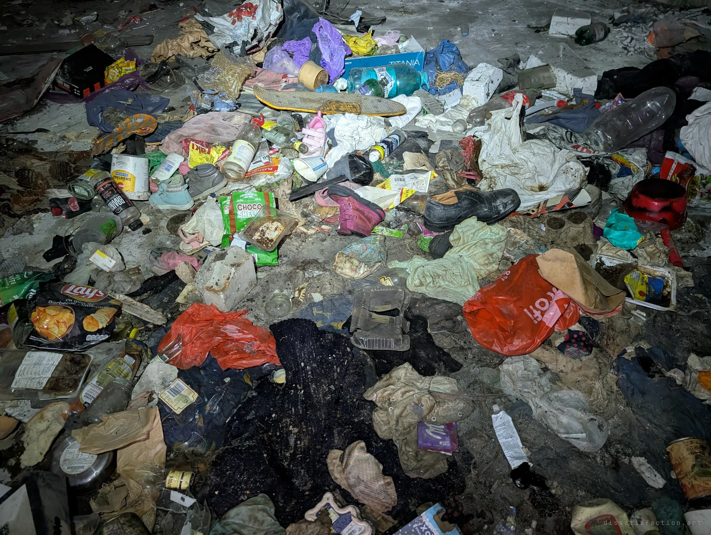 A floor buried under rubbish: food packets, bottles, children's shoes, a skateboard and rotting clothes, lit by a flashlight. The small shoes make the mess feel more personal.