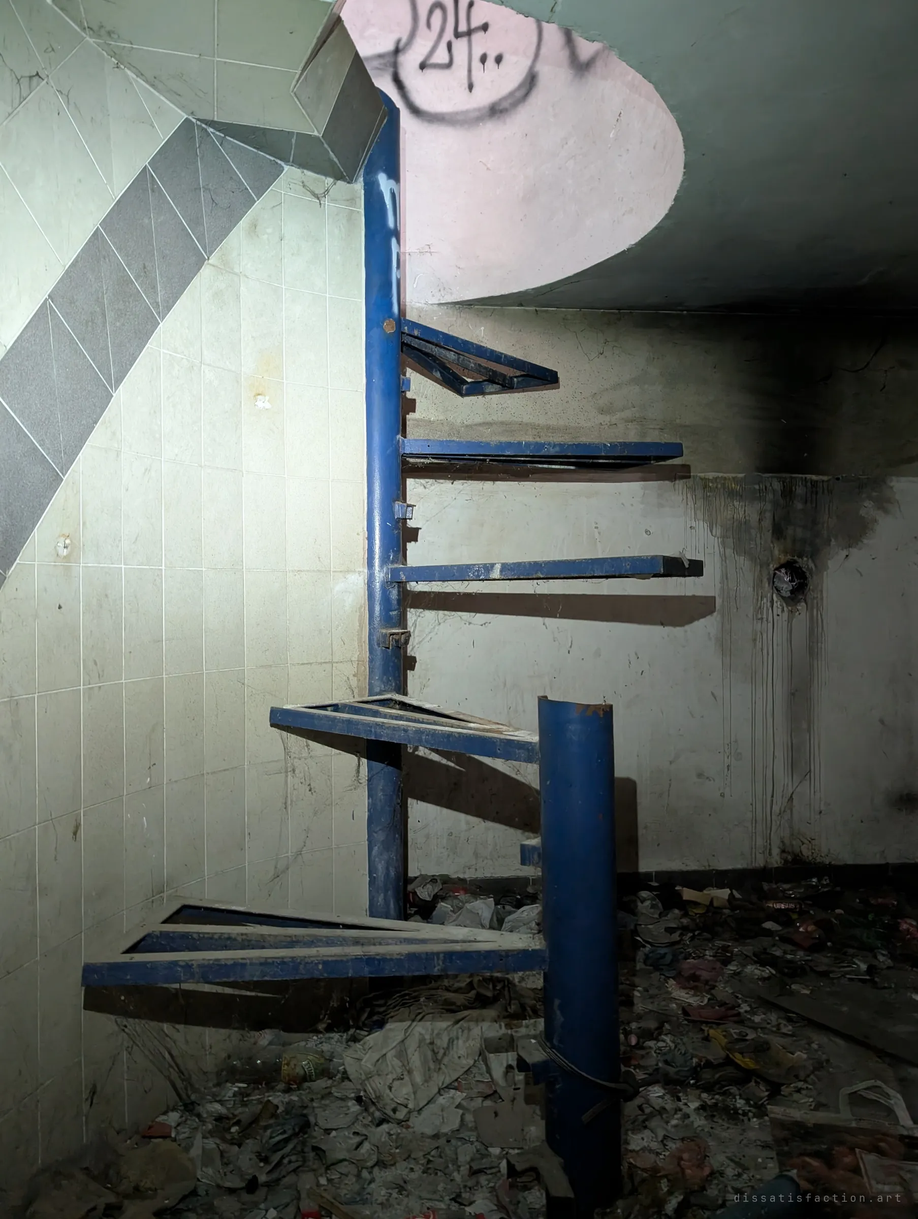 A blue steel spiral staircase with missing and bent treads rises to a hole in the ceiling above a soot-stained wall. Most of the stairs are gone, so the upper floor is cut off.