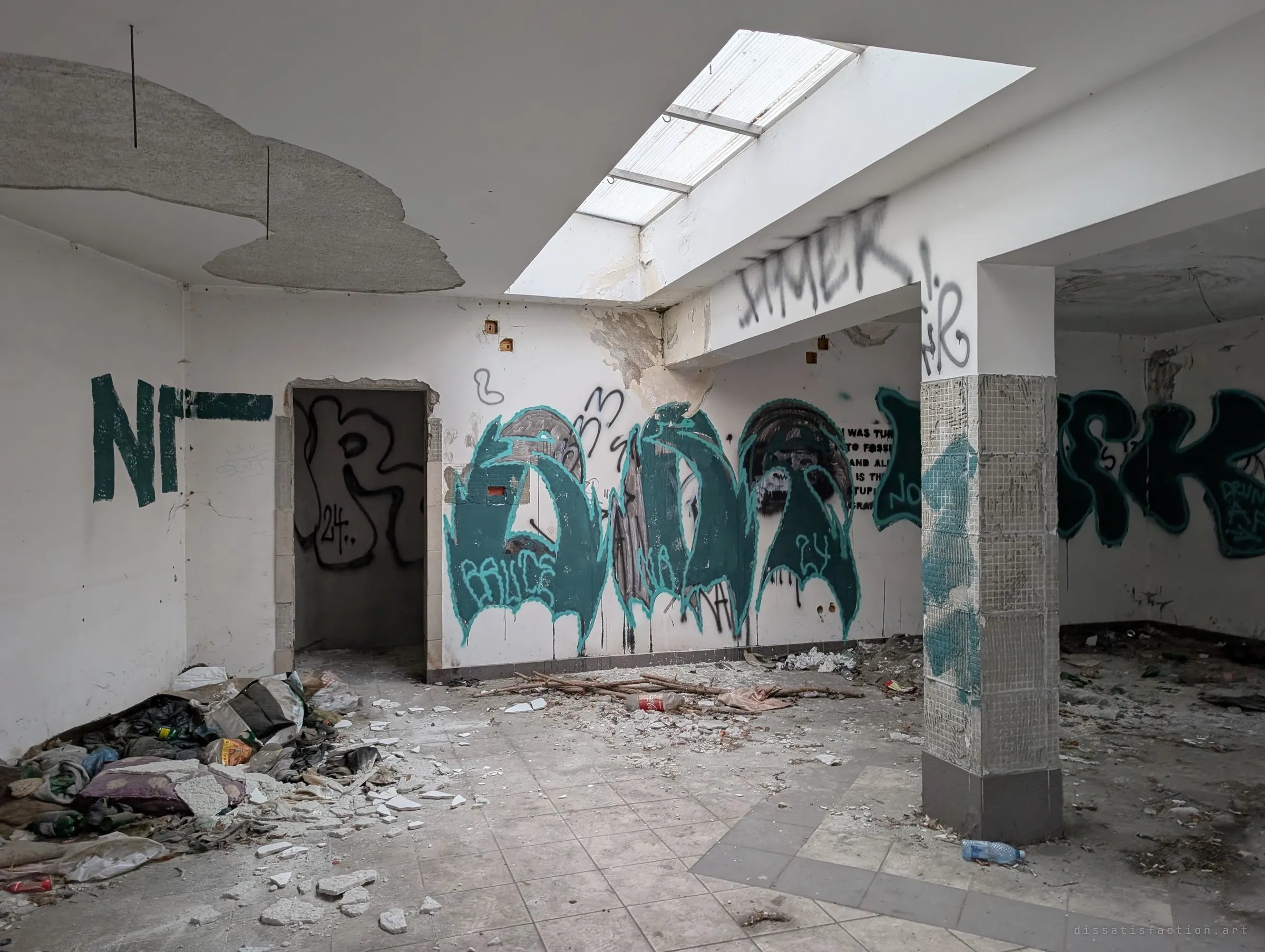 A former shop floor under a skylight, its walls covered in large teal and black graffiti and the ceiling panels falling away. Rubble and a sleeping pile sit in the corner.