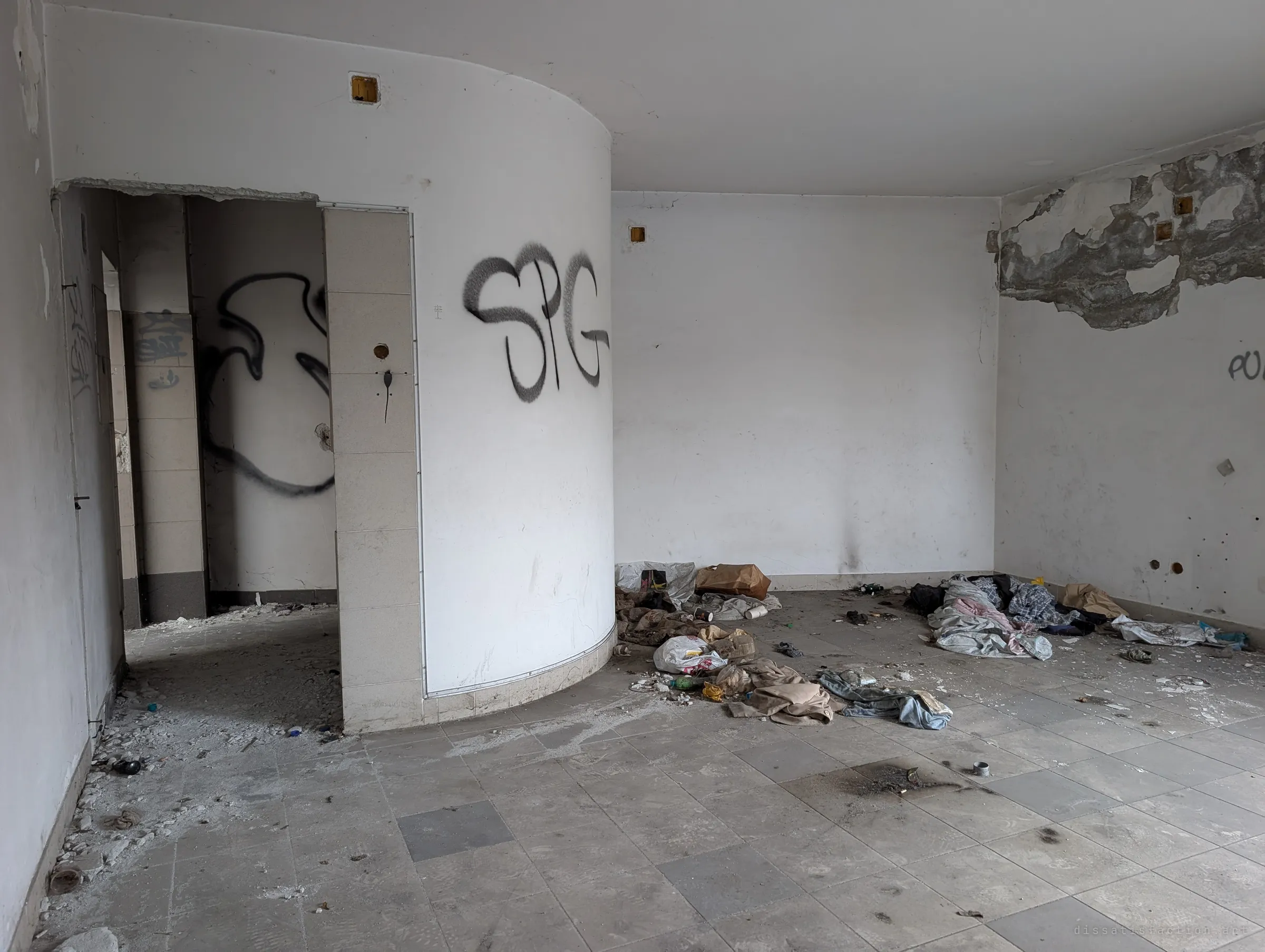 An empty sales room with a curved white wall tagged in black spray paint, a scorch mark on the tiles and clothes dumped along the back wall. It shows how bare the building is now.