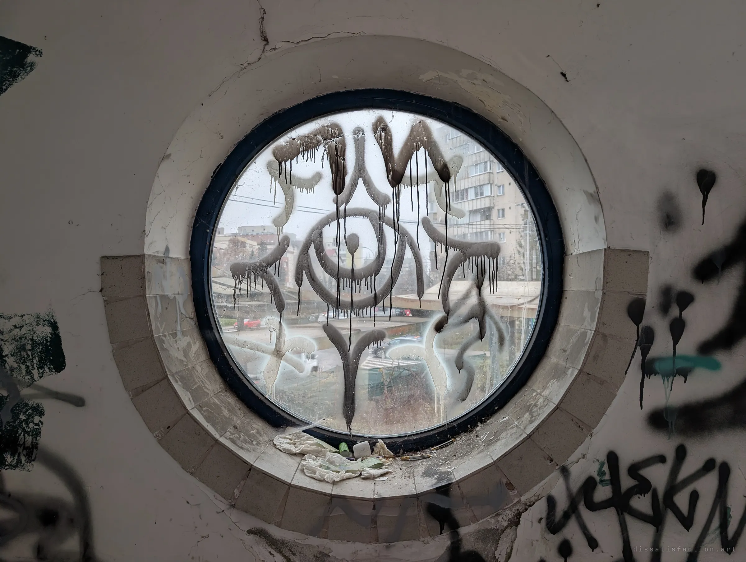 A round porthole window sprayed with a dripping graffiti eye, with grey city blocks faintly visible through the glass. The eye seems to stare back at whoever is inside.