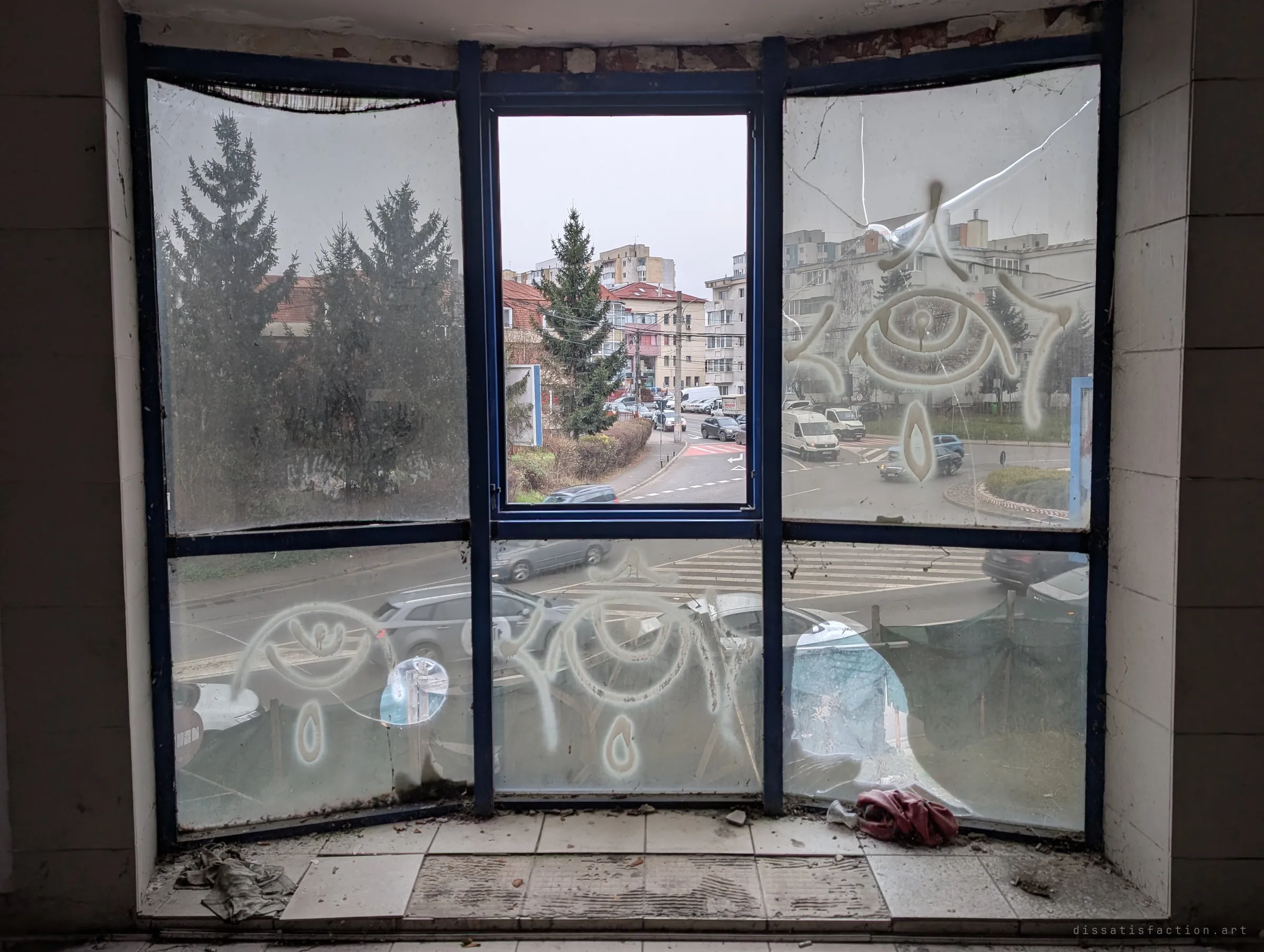 A cracked, graffiti-painted shopfront window looking out on busy traffic under a grey sky. The eye tags on the glass make the empty room feel watched.