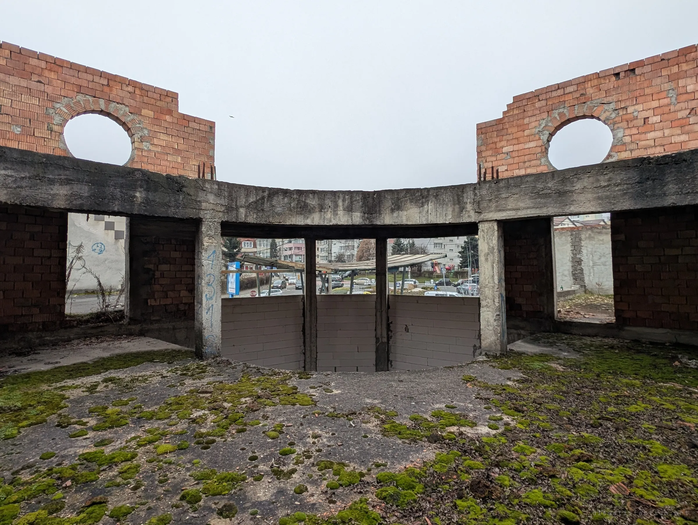 A curved, roofless concrete ring with circular brick openings, its window bays looking straight out onto busy city traffic. Moss has taken the floor while the street carries on just beyond the unfinished walls.