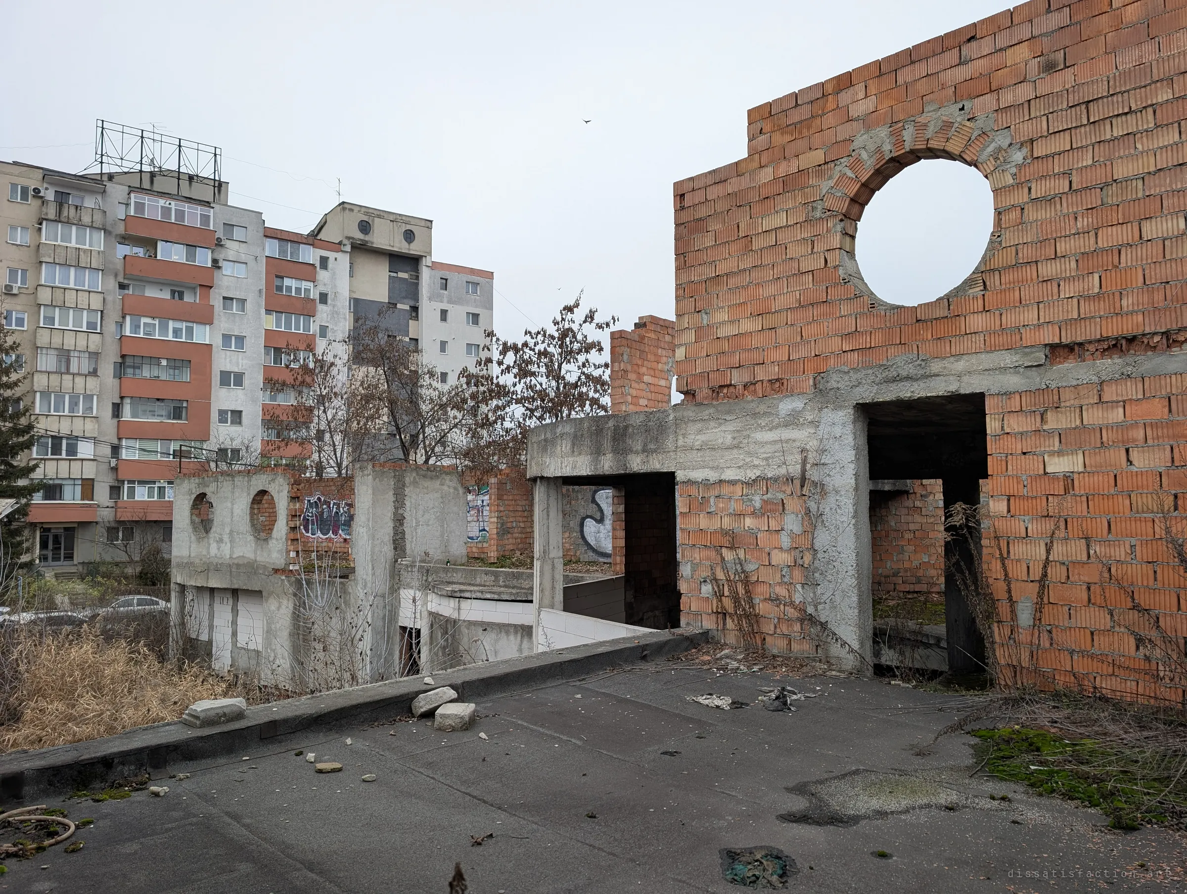The upper deck of the unfinished building, brick walls with a round porthole opening rising beside tall apartment blocks. The ruin sits wedged into a lived-in neighbourhood.