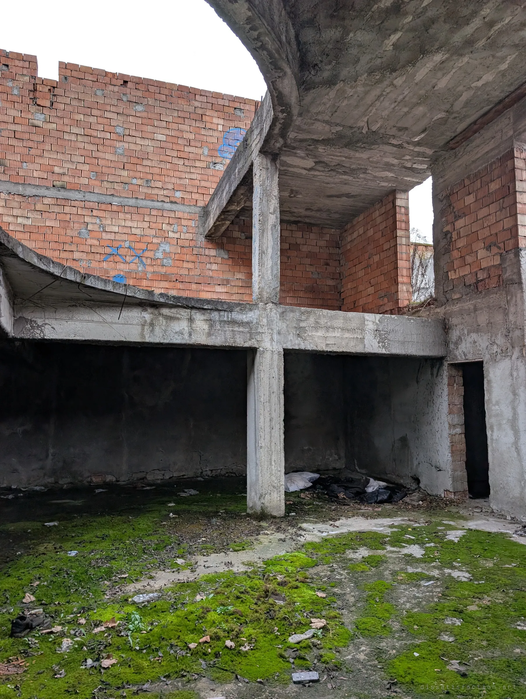 A curved concrete slab sweeps over a moss-covered floor, with a dark doorway and a pile of bags tucked under the overhang. The shape shows the building's odd round plan.
